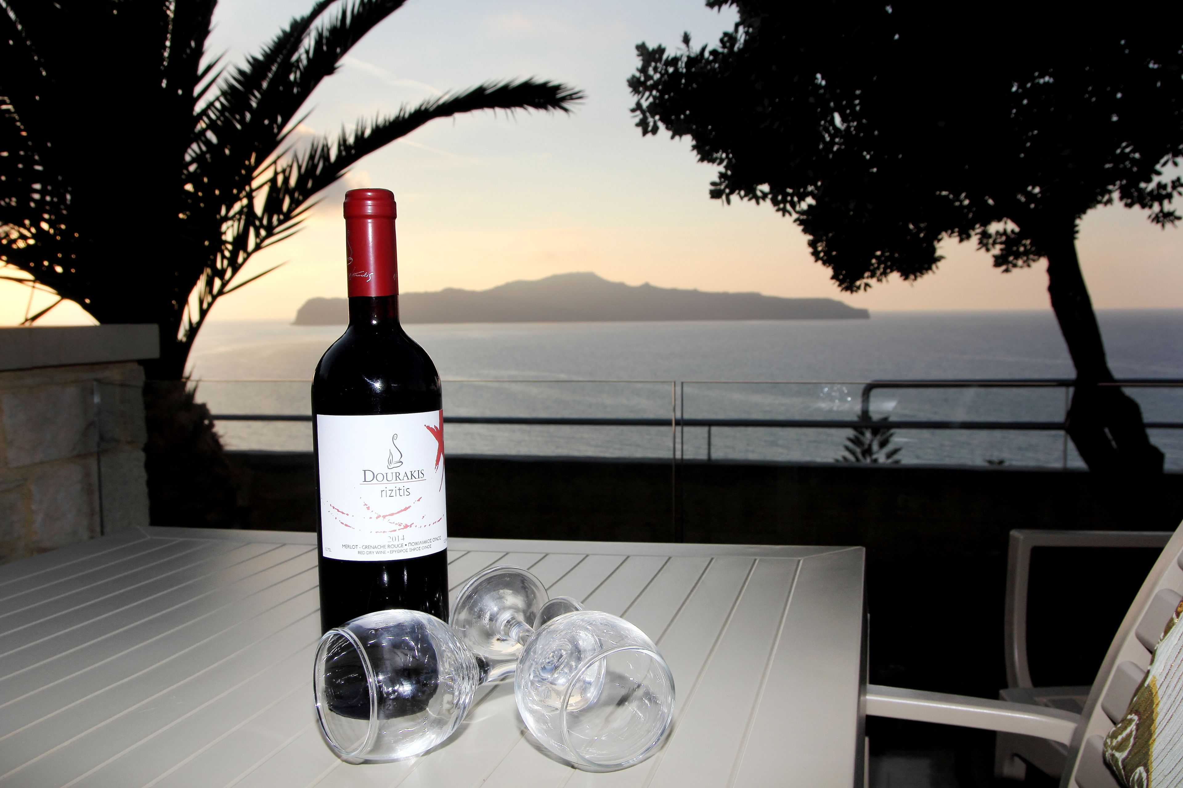 A bottle of Doux Rizzis wine and three empty wine glasses on a white table with a view of the sea and mountains in the background.
