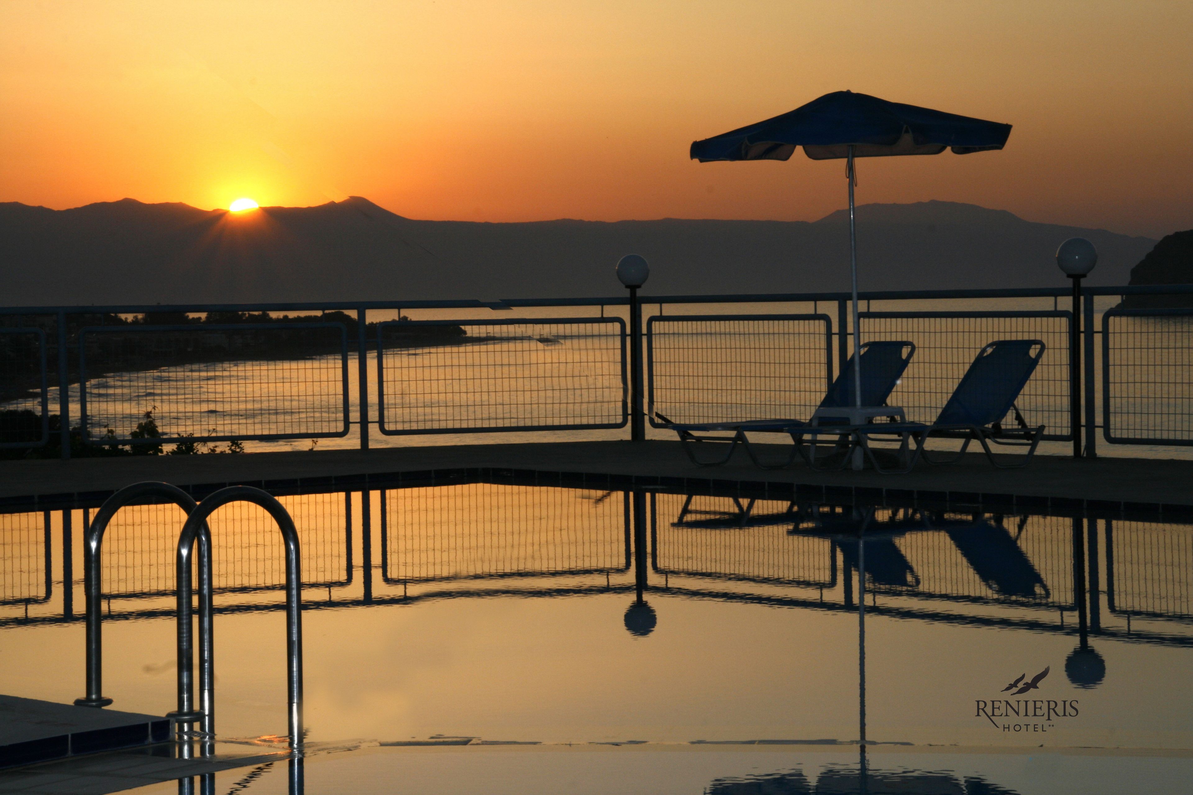 A poolside view with lounge chairs and an umbrella, reflecting the sunset over a body of water with mountains in the background.