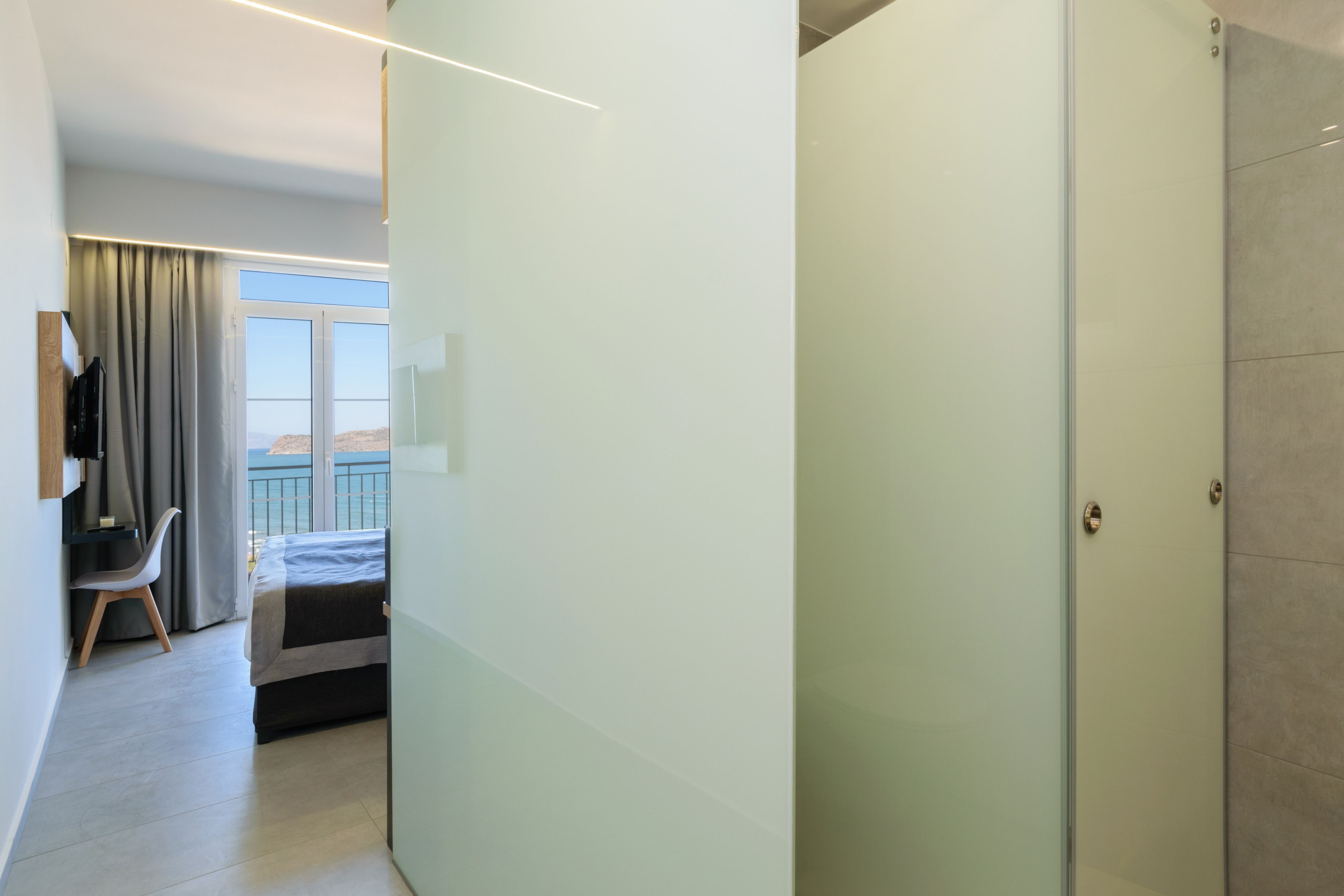 Habitació d'hotel moderna amb un llit, una taula de treball i una cadira. Hi ha una finestra gran amb cortines, i una porta que dona a una altra habitació.
