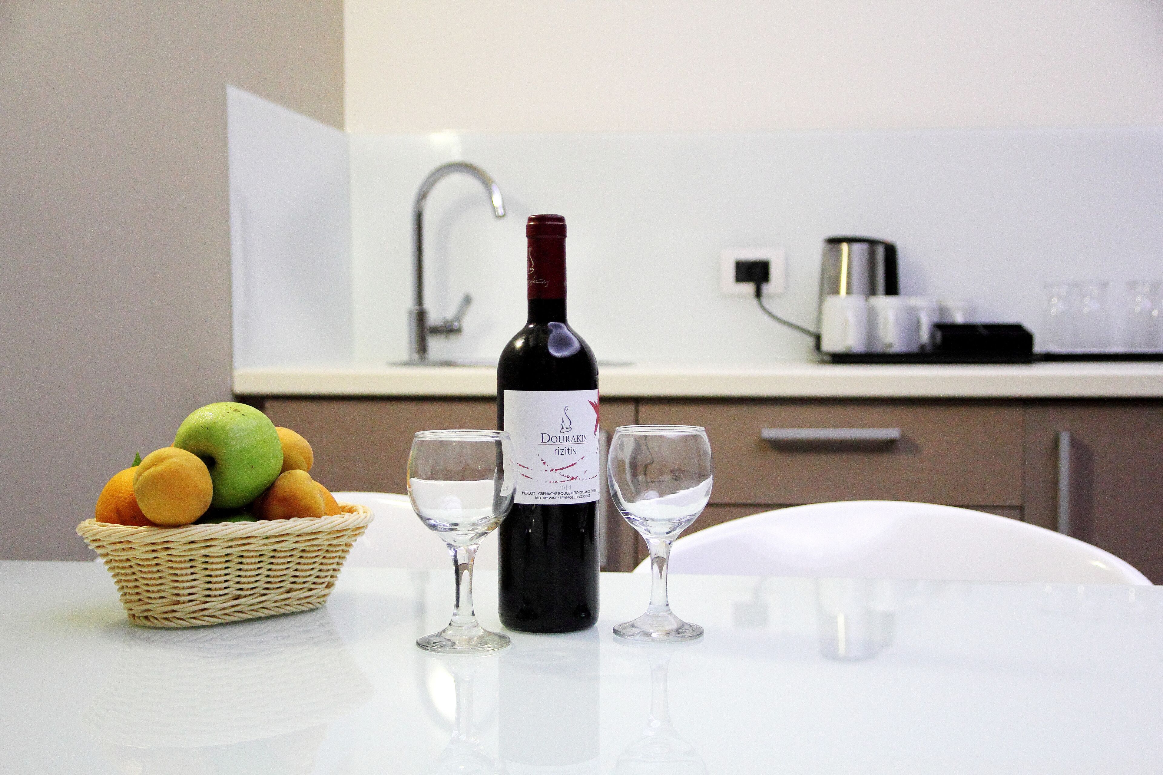 A bottle of wine, two glasses, and a basket of fruit on a table in a modern kitchen.