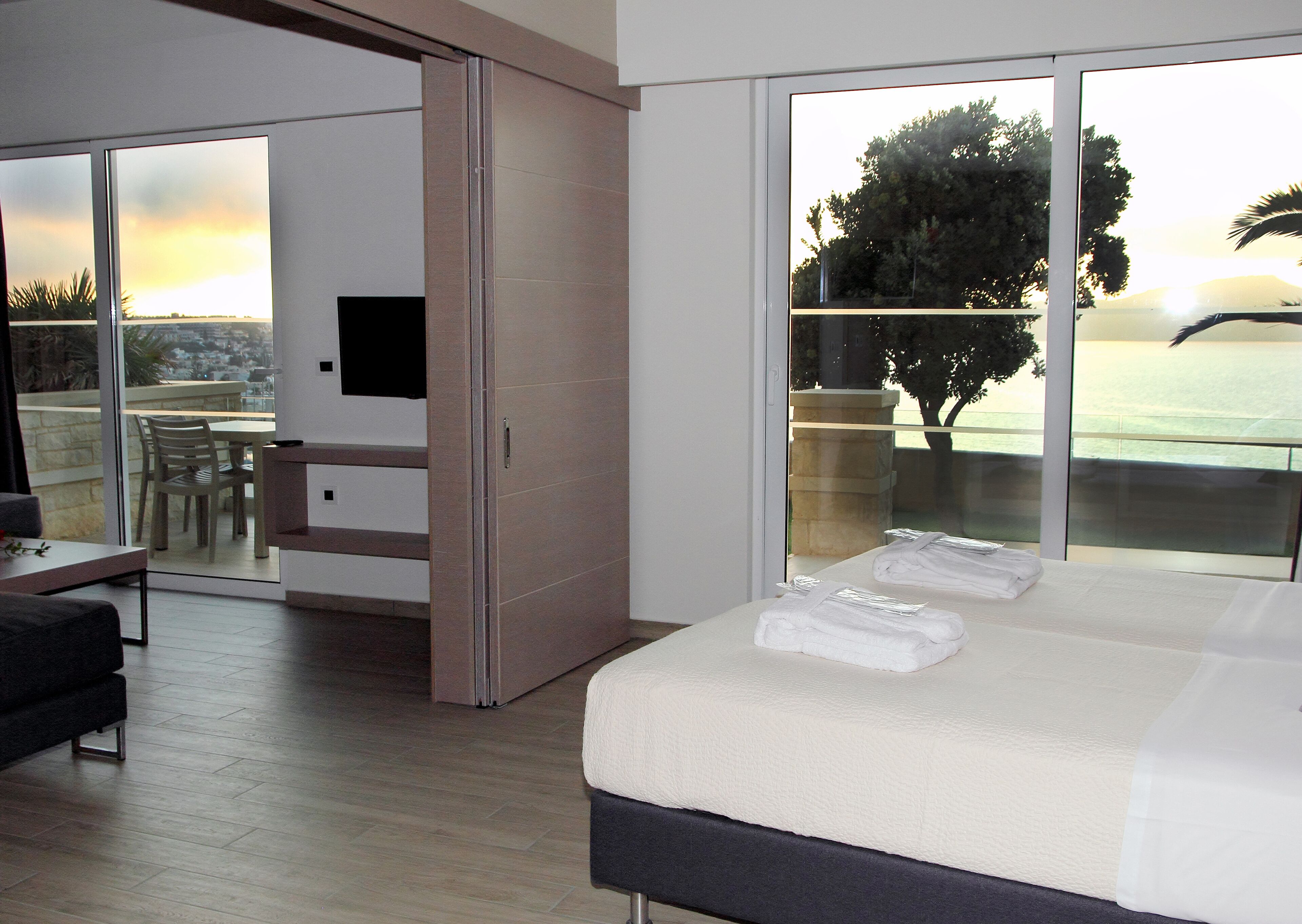A modern hotel room with a large bed, a TV, a balcony with a view of the sea, and a dining area.