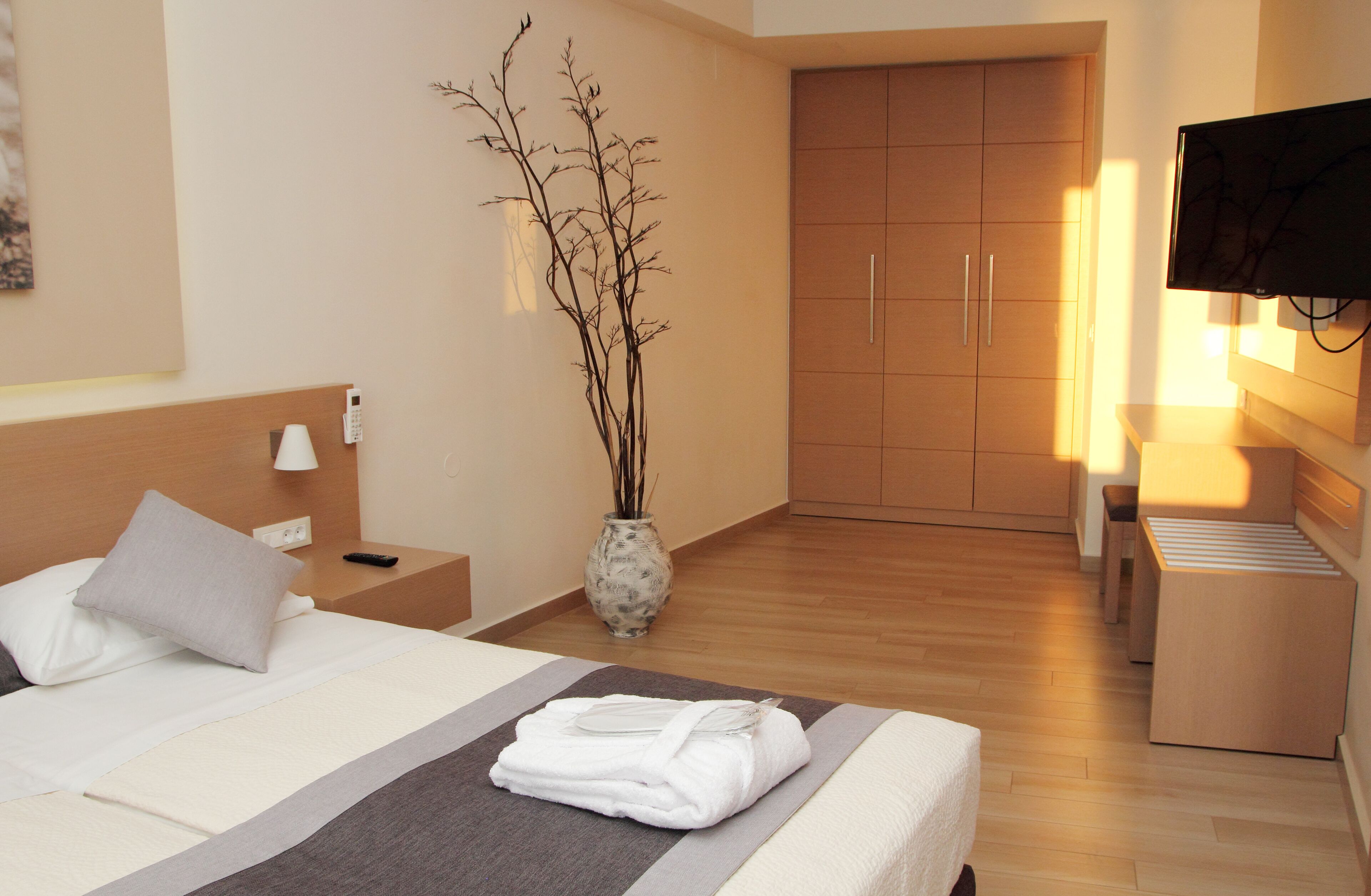 Une chambre d’hôtel comprenant un lit, une table de chevet, une lampe, un vase avec des branches, une armoire et un téléviseur.