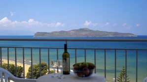 Een balkon met een tafel waarop een wijnfles en een schaal fruit staan, met uitzicht op een strand en een eiland in de verte.