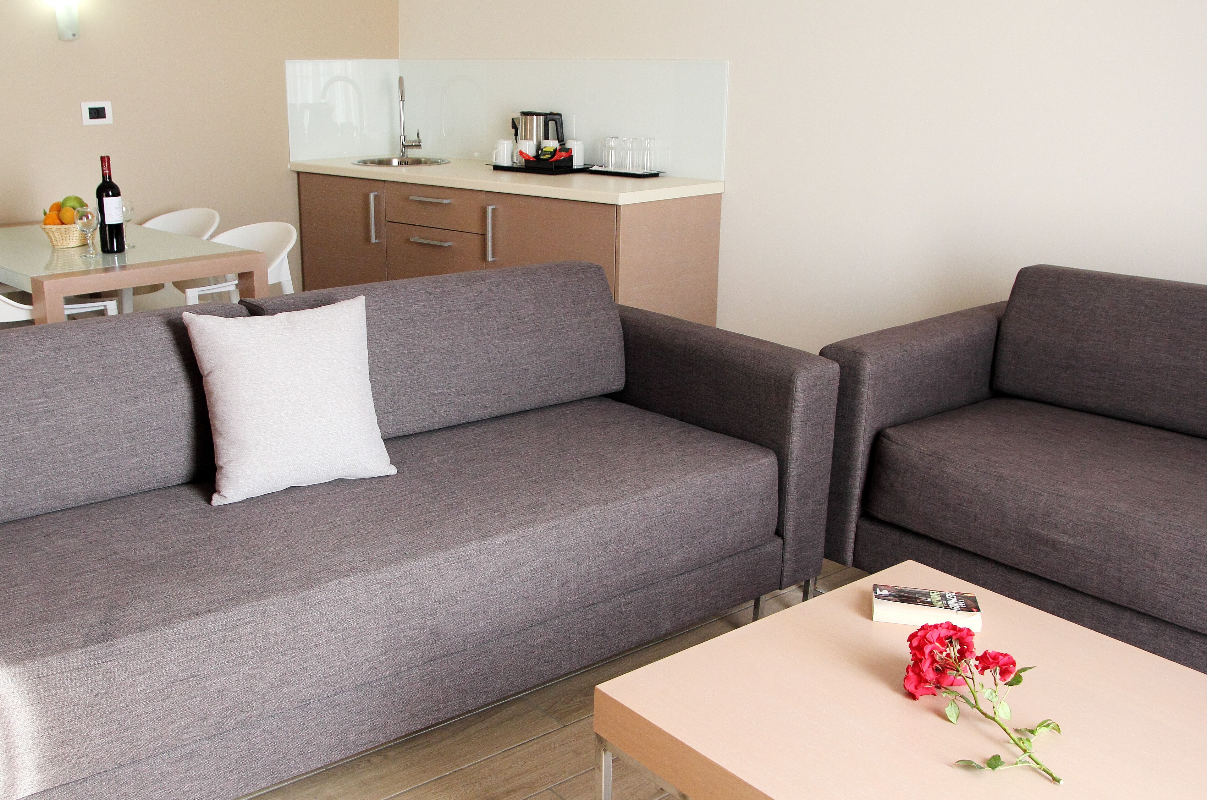 A modern living room with two grey sofas, a small table with a red flower, and a kitchen area in the background.