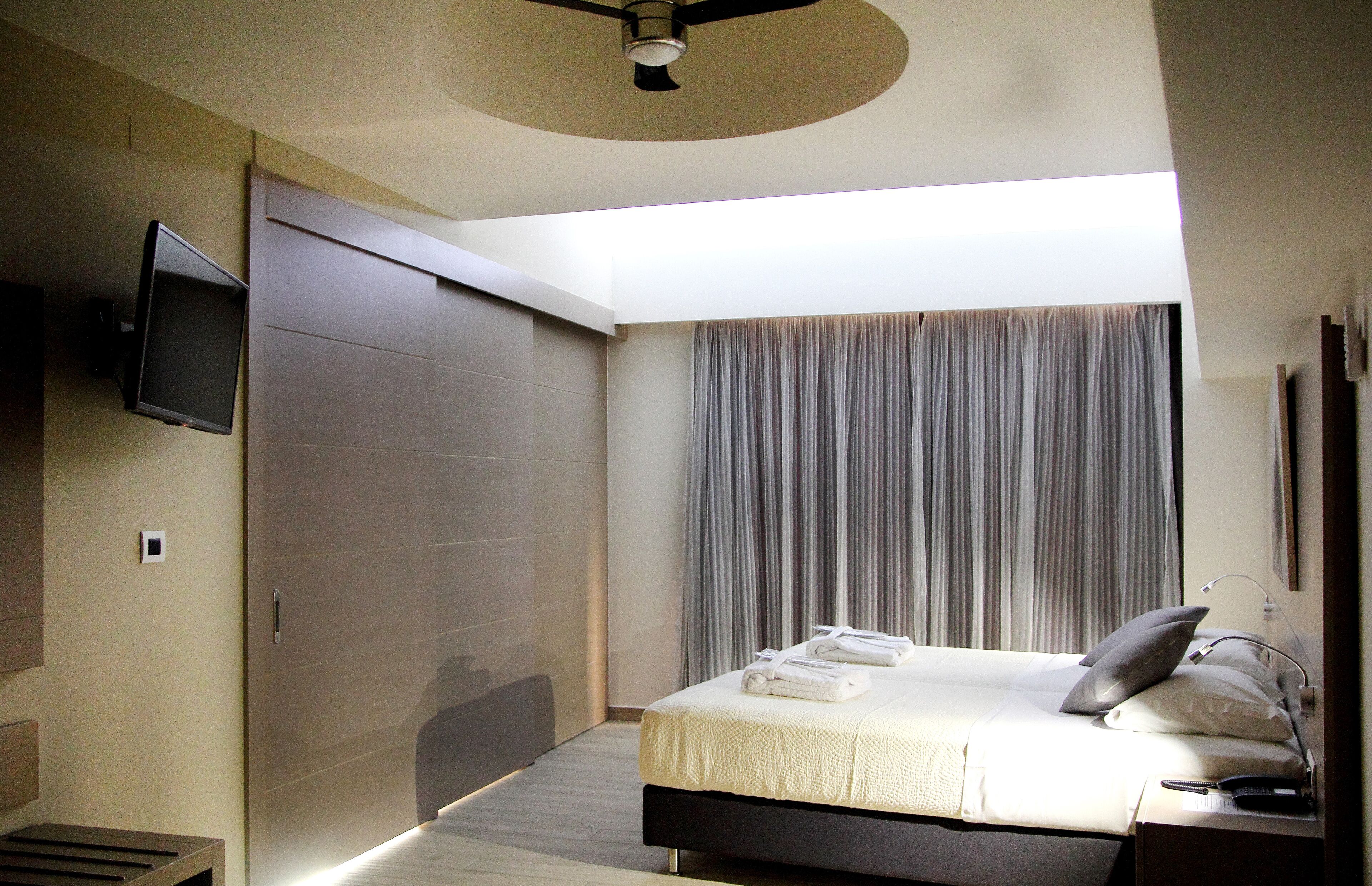 Habitación de hotel moderna con una cama, un televisor de pantalla plana montado en la pared, un ventilador de techo y un ventanal con cortinas.