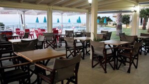 Een overdekt terras met tafels en stoelen, met uitzicht op een zwembad en strand.