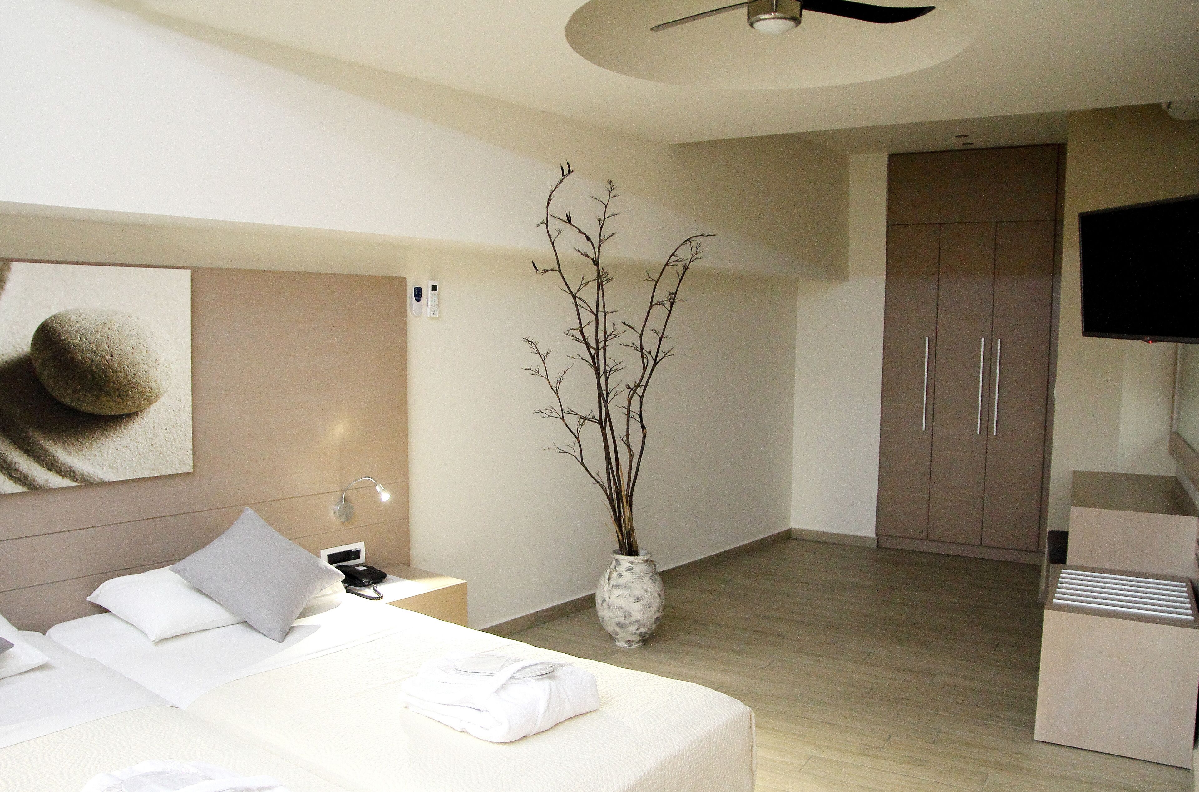 A modern hotel room with a bed, bedside table, lamp, vase with branches, wardrobe, and a television.