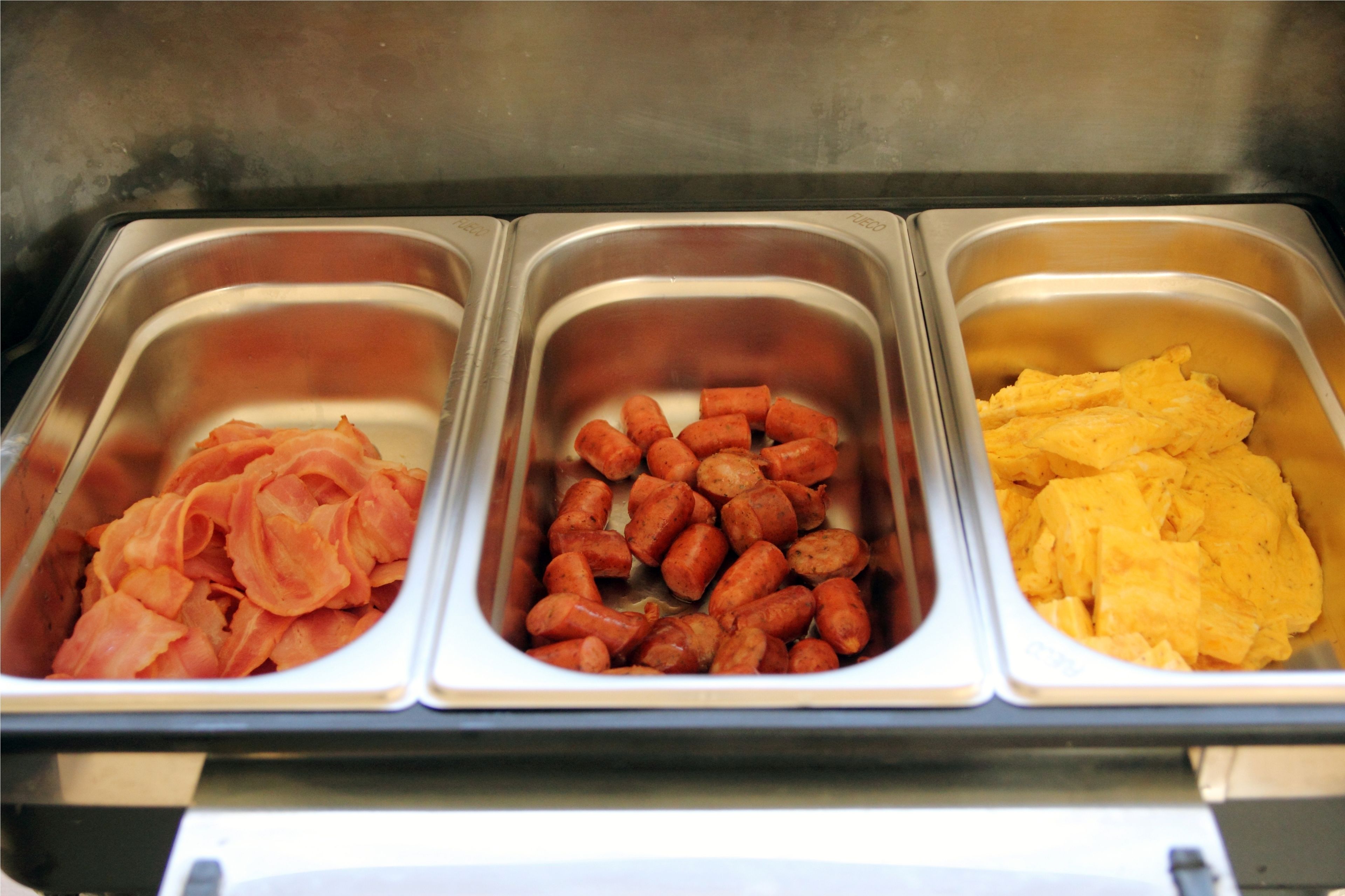 Three stainless steel food dispensers with different food items: sliced ham, cooked sausages, and scrambled eggs.