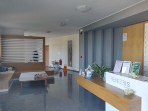 Zona de recepción de un hotel con un mostrador de madera, un área de espera con un sofá y una mesa de centro, y un extintor de pared.