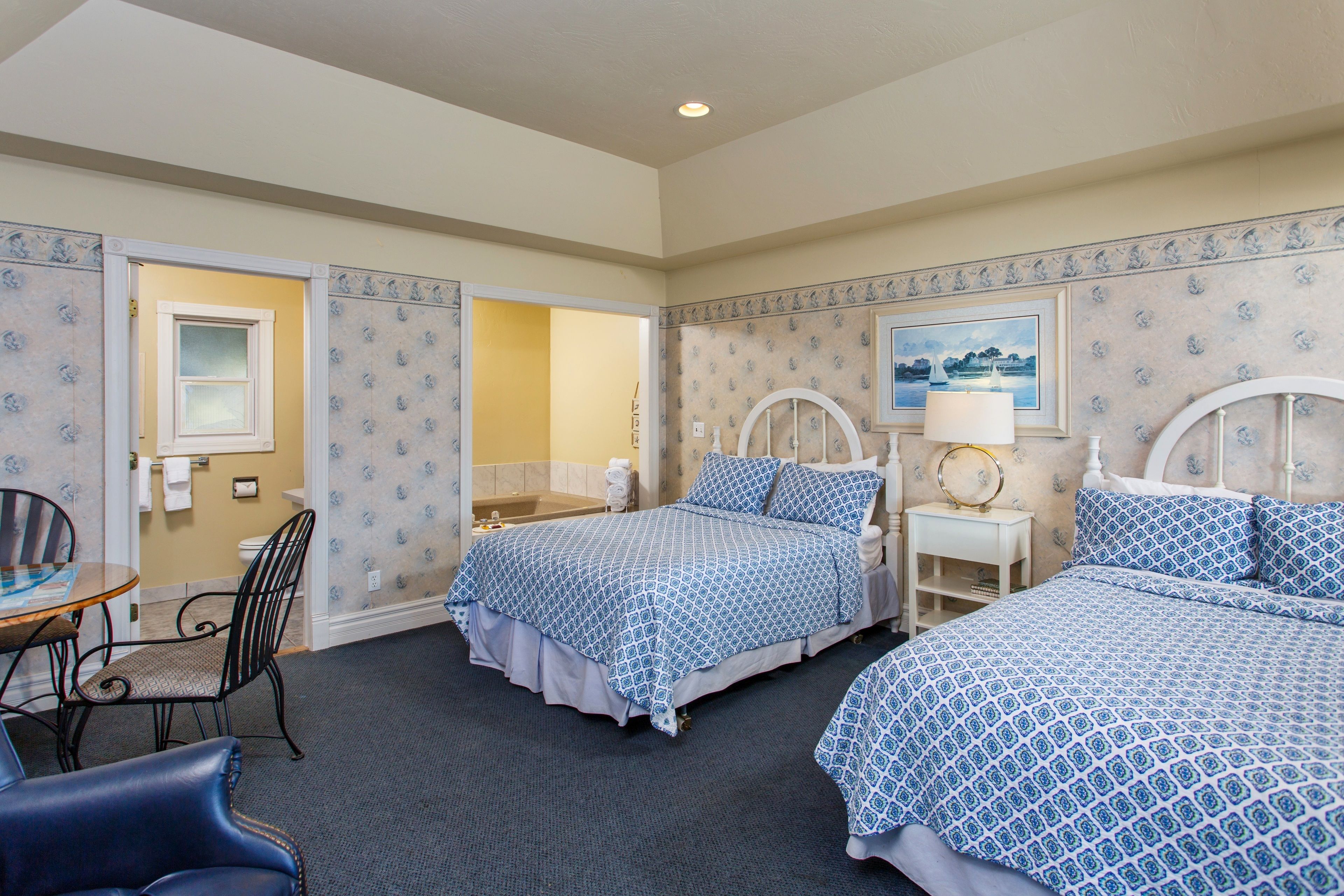 A hotel room with two beds, a sitting area with a table and chairs, a bathroom with a bathtub, and a painting on the wall.