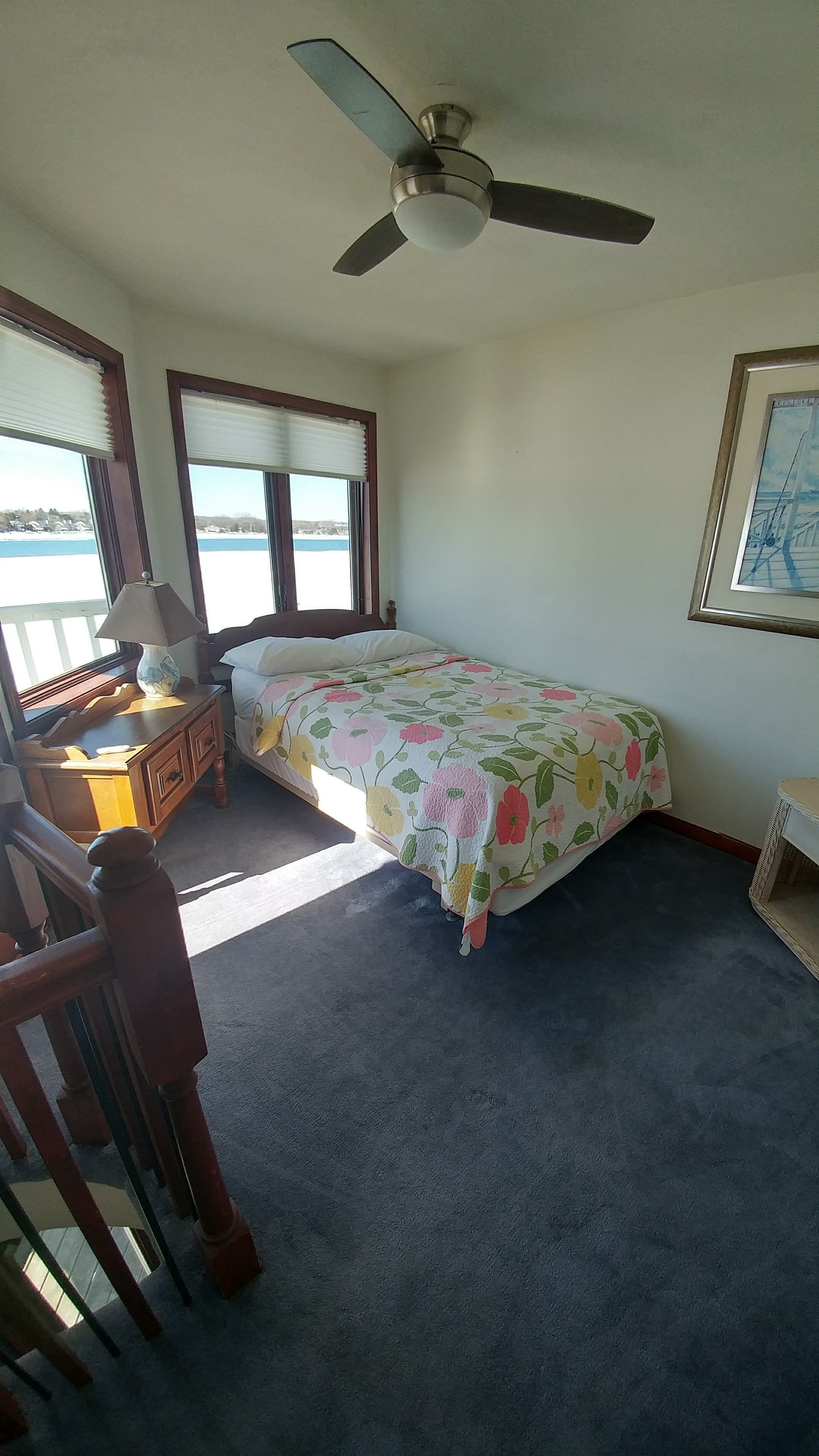 A bedroom with a bed, a desk, a chair, a ceiling fan, and a window with a view of the water.