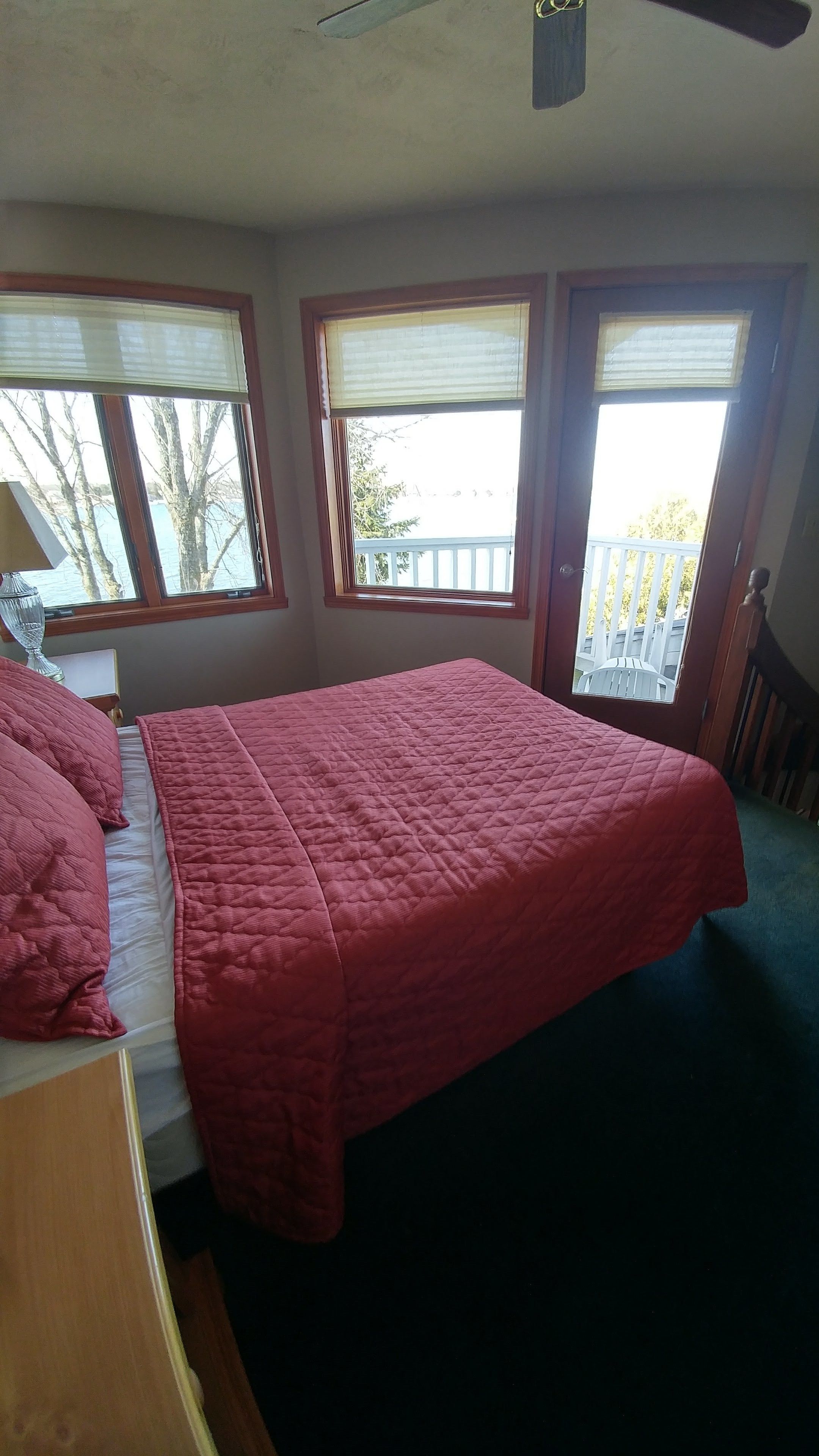 A bedroom with a large bed, two windows with blinds, a ceiling fan, and a view of a balcony.