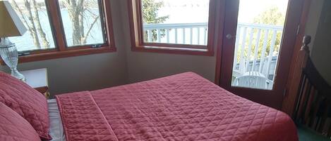 A bedroom with a large bed, two windows with blinds, a ceiling fan, and a view of a balcony.