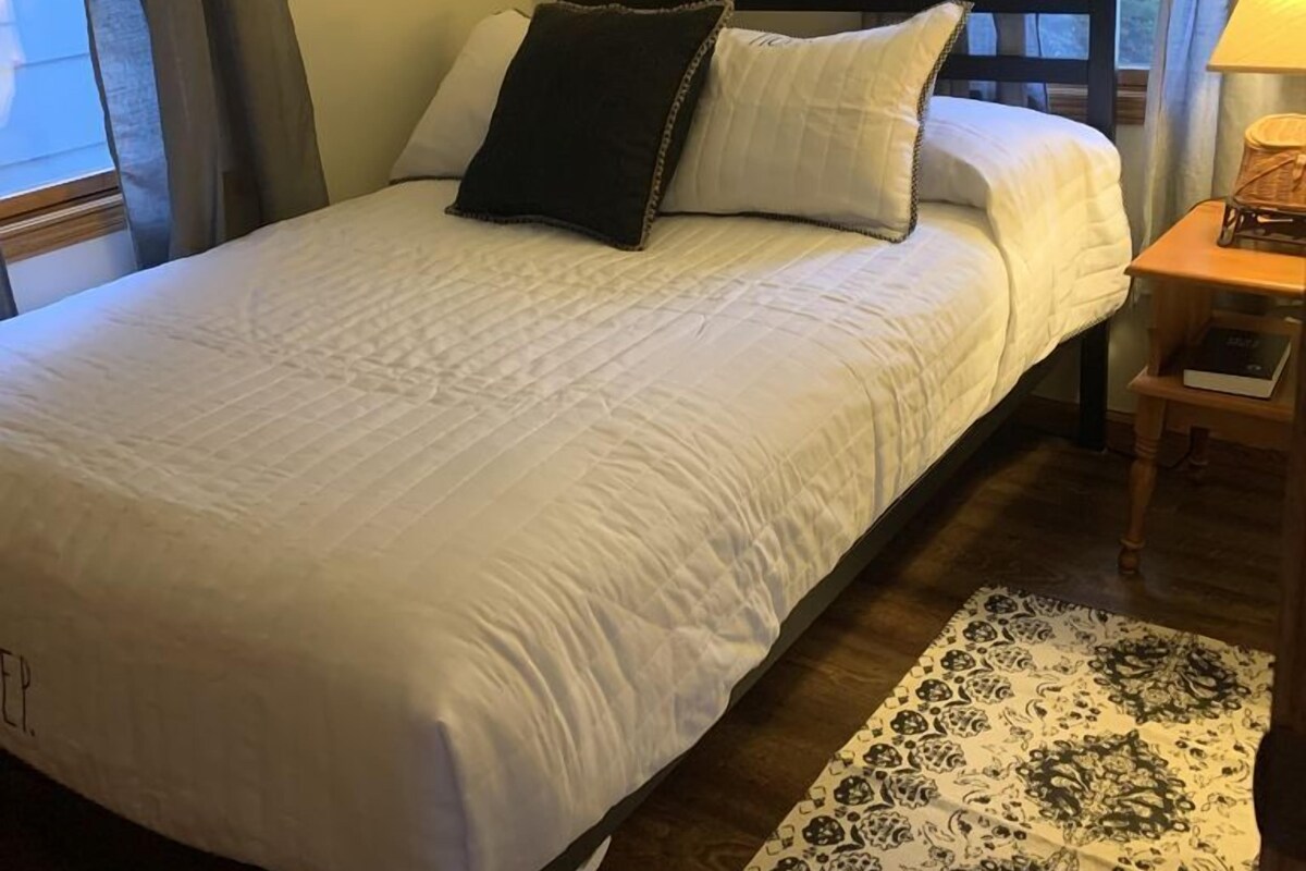 A neatly made bed with white bedding and two pillows, a bedside table with a lamp, and a patterned rug on the floor.