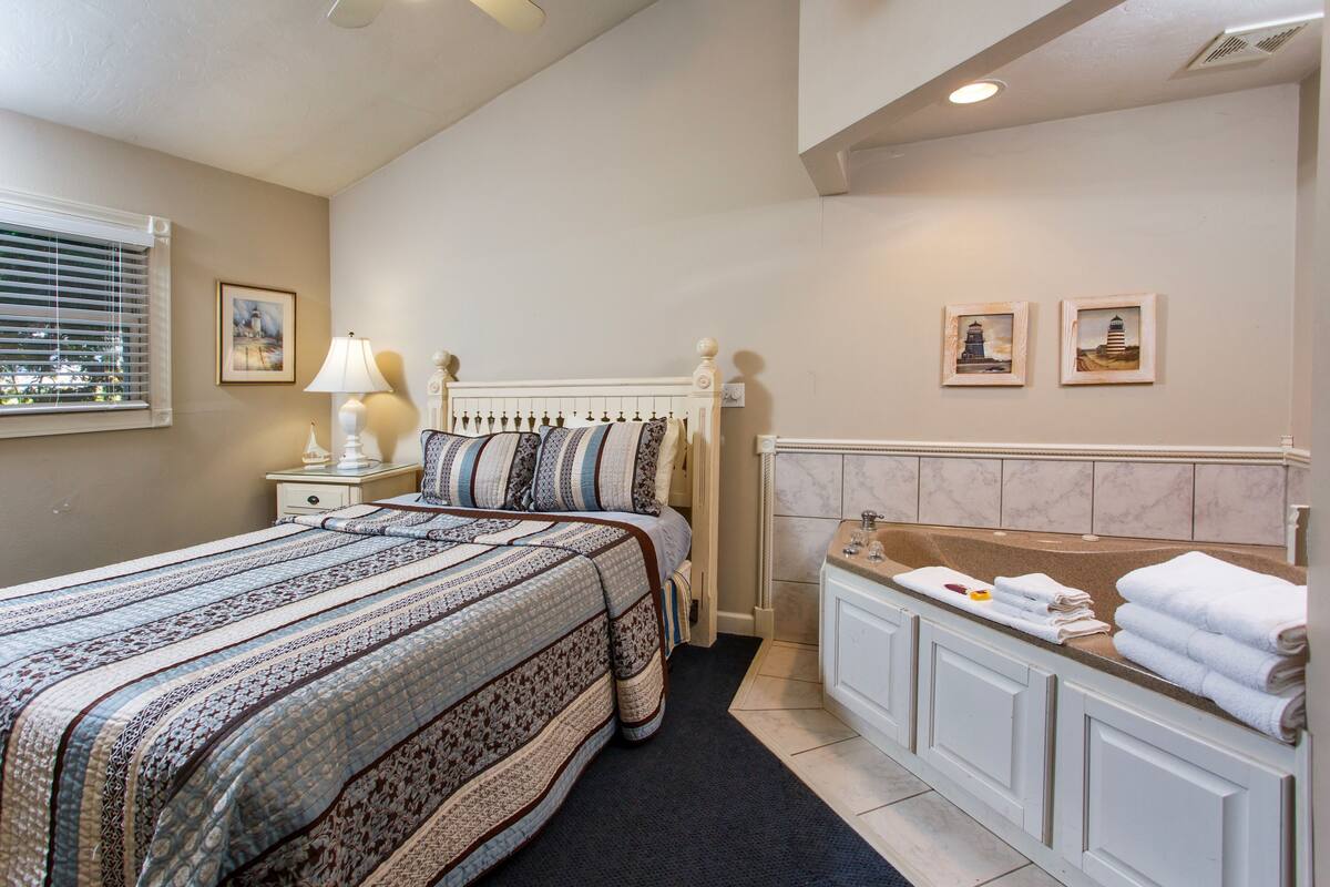 A bedroom with a large bed, a nightstand, a lamp, a window with blinds, and a bathroom with a sink and towels.