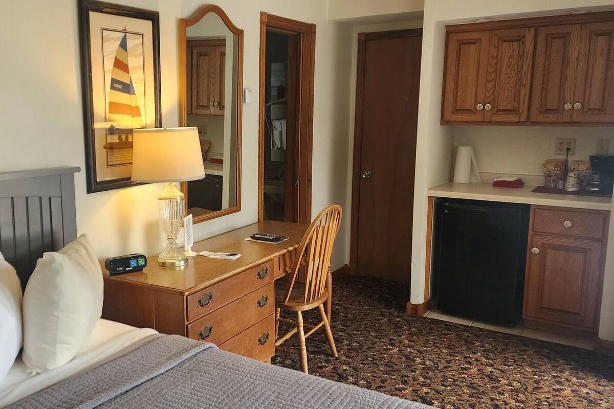 A hotel room with a bed, a desk, a chair, a dresser, a mirror, and a kitchenette.