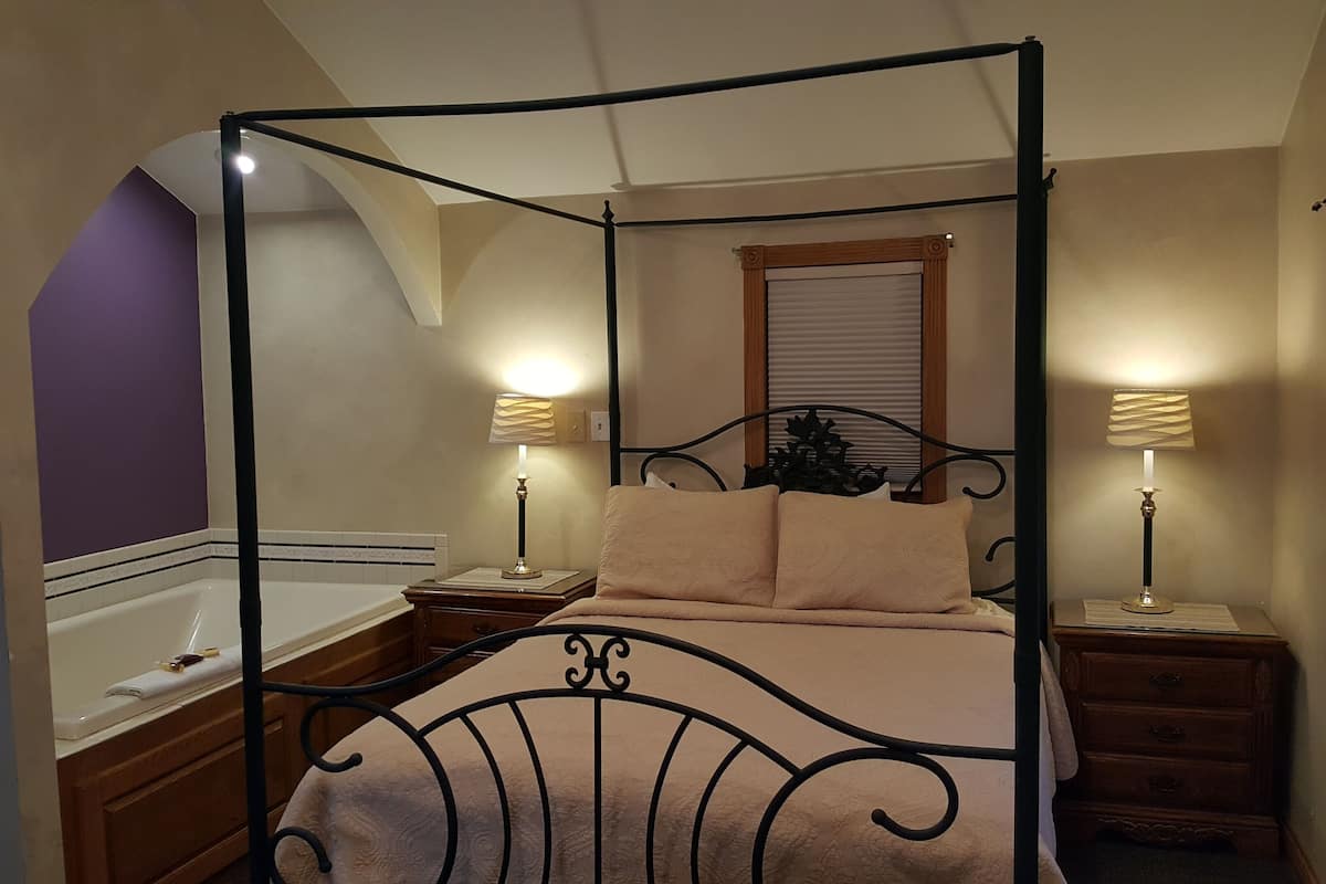 A four-poster canopy bed with a headboard, two bedside tables with lamps, a window with blinds, and a wooden dresser.