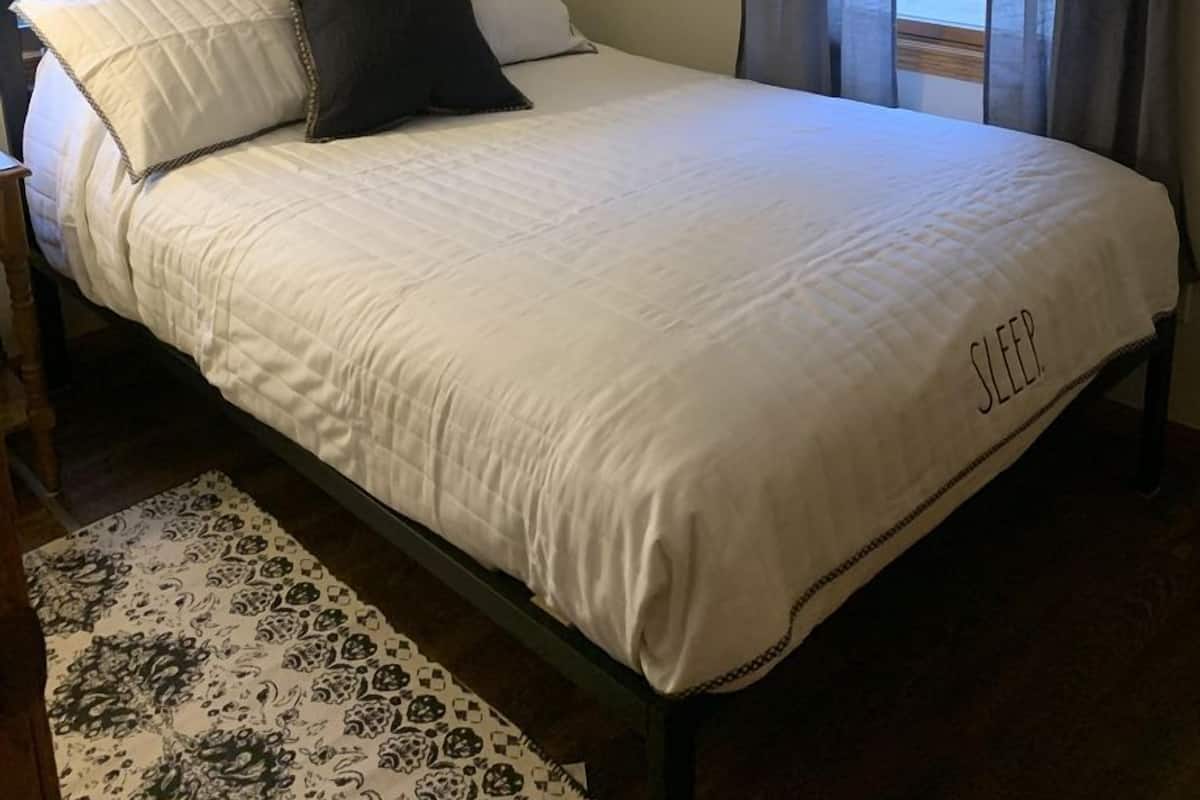 A neatly made bed with a white comforter and pillows, a black metal frame, a hanging light fixture, and a patterned rug.