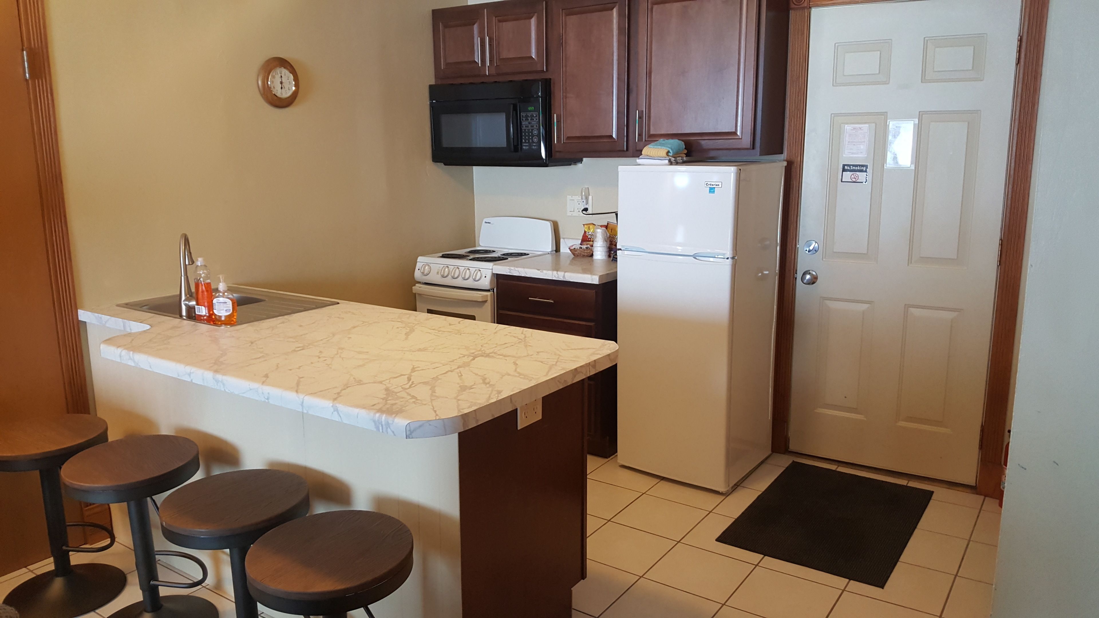 A compact kitchen with a white marble countertop, a stove, a microwave, and a refrigerator. There are two stools at the counter and a clock on the wall.