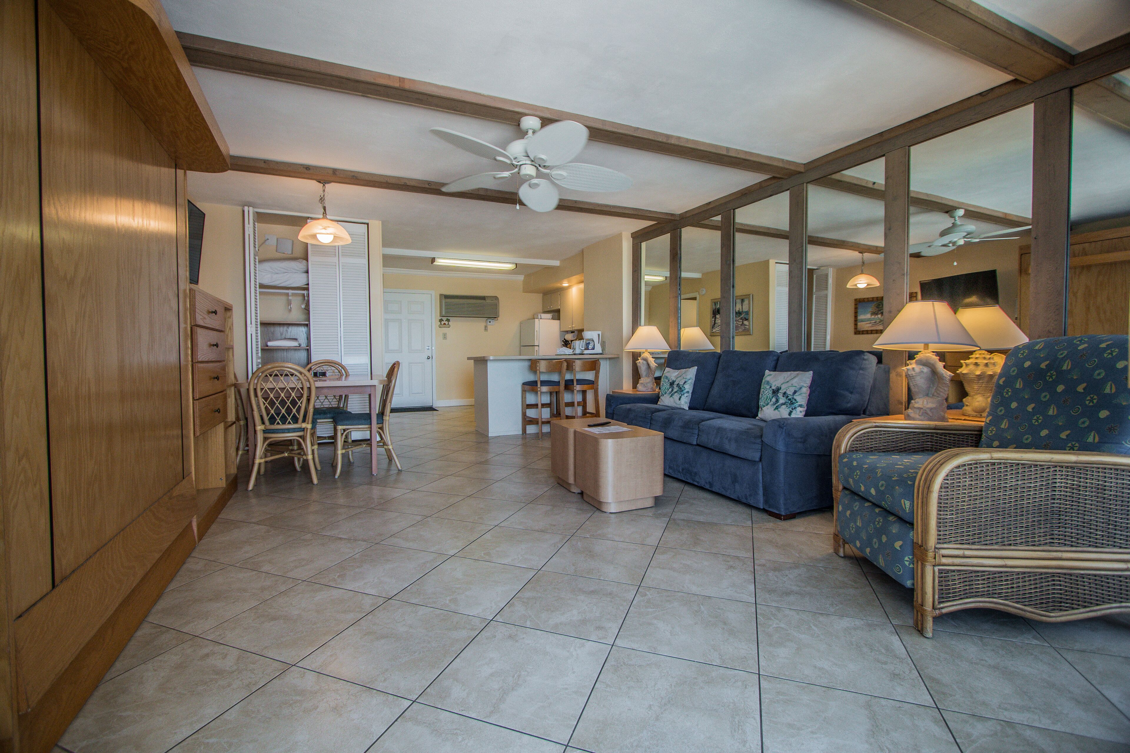 A spacious living area with a sofa, chairs, and a dining table. There is a kitchen in the background with a refrigerator and a microwave.