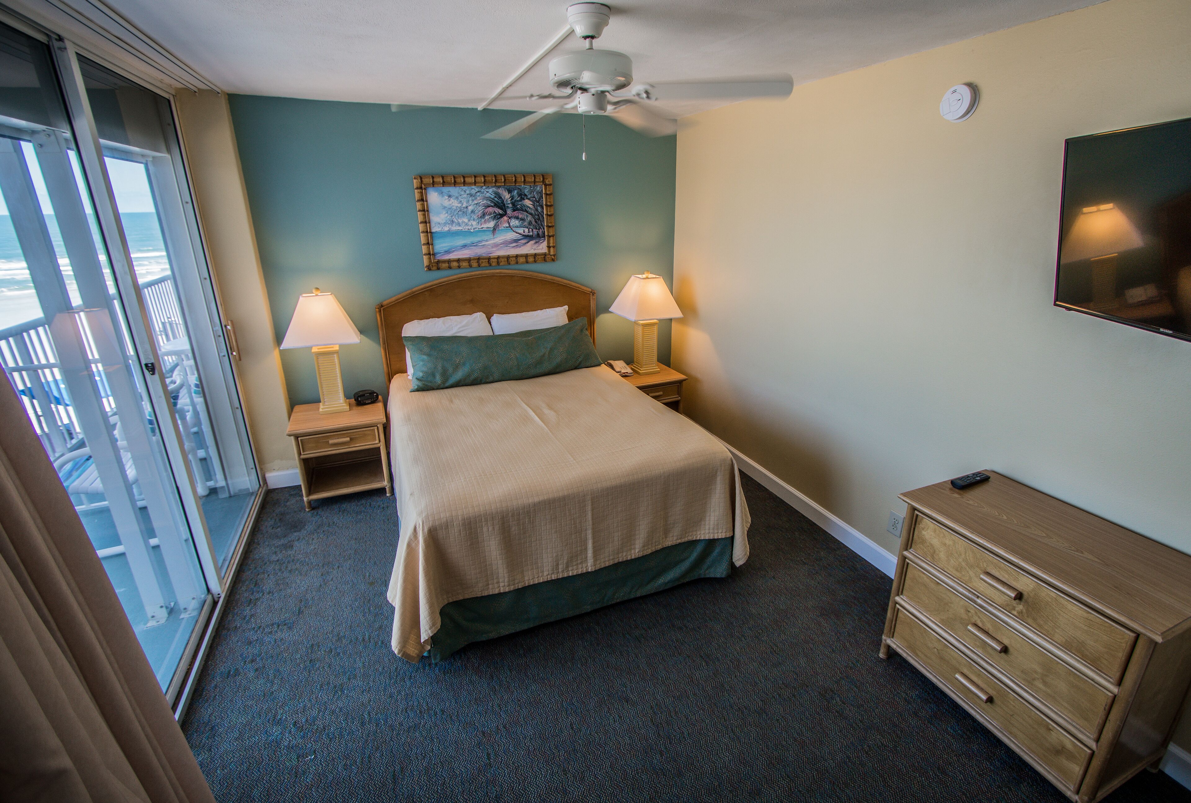 A hotel room with a bed, two bedside lamps, a ceiling fan, a TV, a wooden dresser, and a balcony with a view of the ocean.