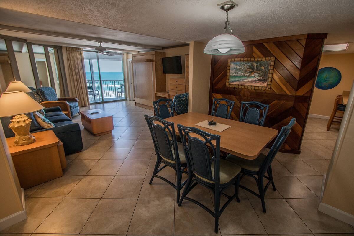 A spacious living area with a dining table, chairs, and a sofa. There is a balcony with a view of the ocean.