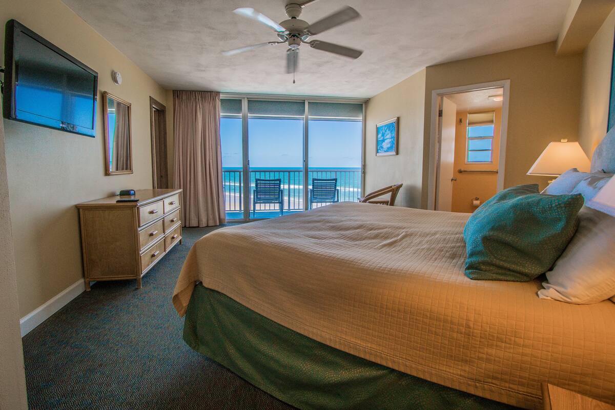 A hotel room with a large bed, a ceiling fan, a television, a dresser, and a view of the ocean through the balcony doors.