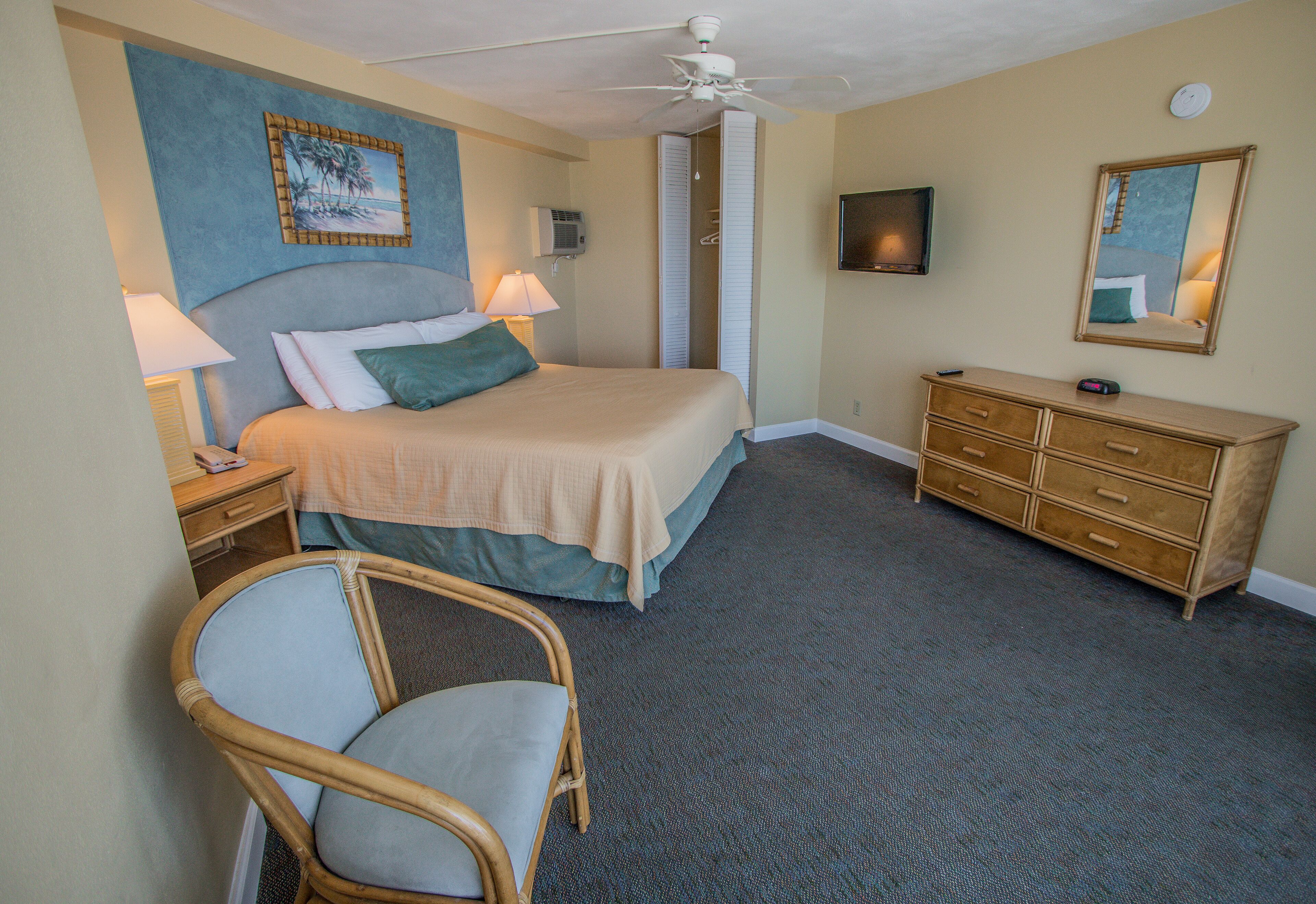 A hotel room with a bed, a chair, a dresser, a ceiling fan, a TV, and a painting on the wall.