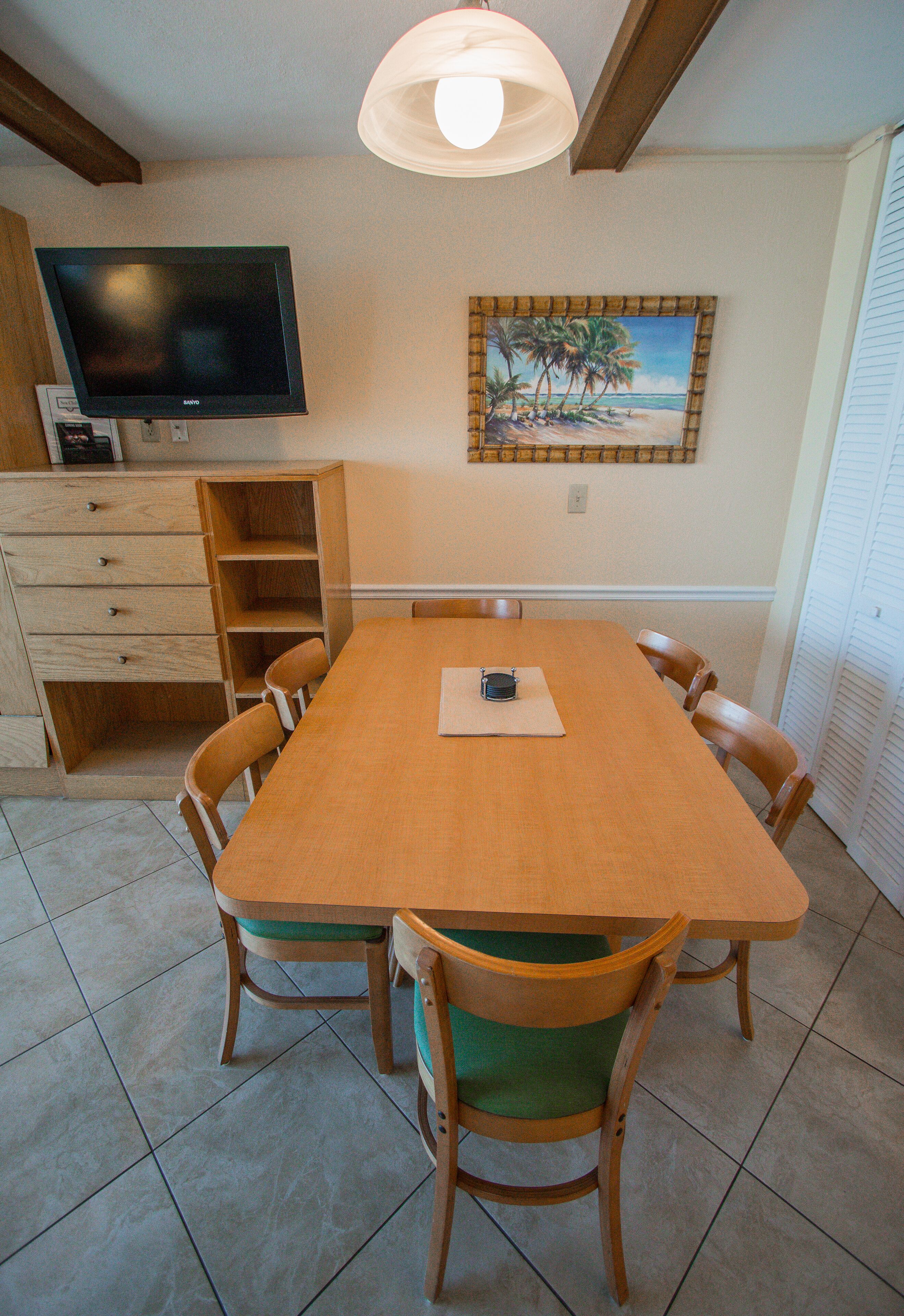 A dining area with a long wooden table, six chairs, a television mounted on the wall, and a painting of a beach scene.