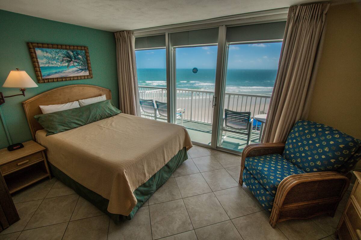 A hotel room with a bed, a chair, a nightstand, a lamp, and a view of the ocean through large windows.