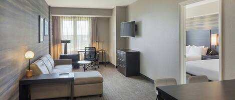 Une chambre d’hôtel équipée d’un canapé, d’un bureau, d’un téléviseur fixé au mur, d’un lit et d’une fenêtre avec des rideaux.