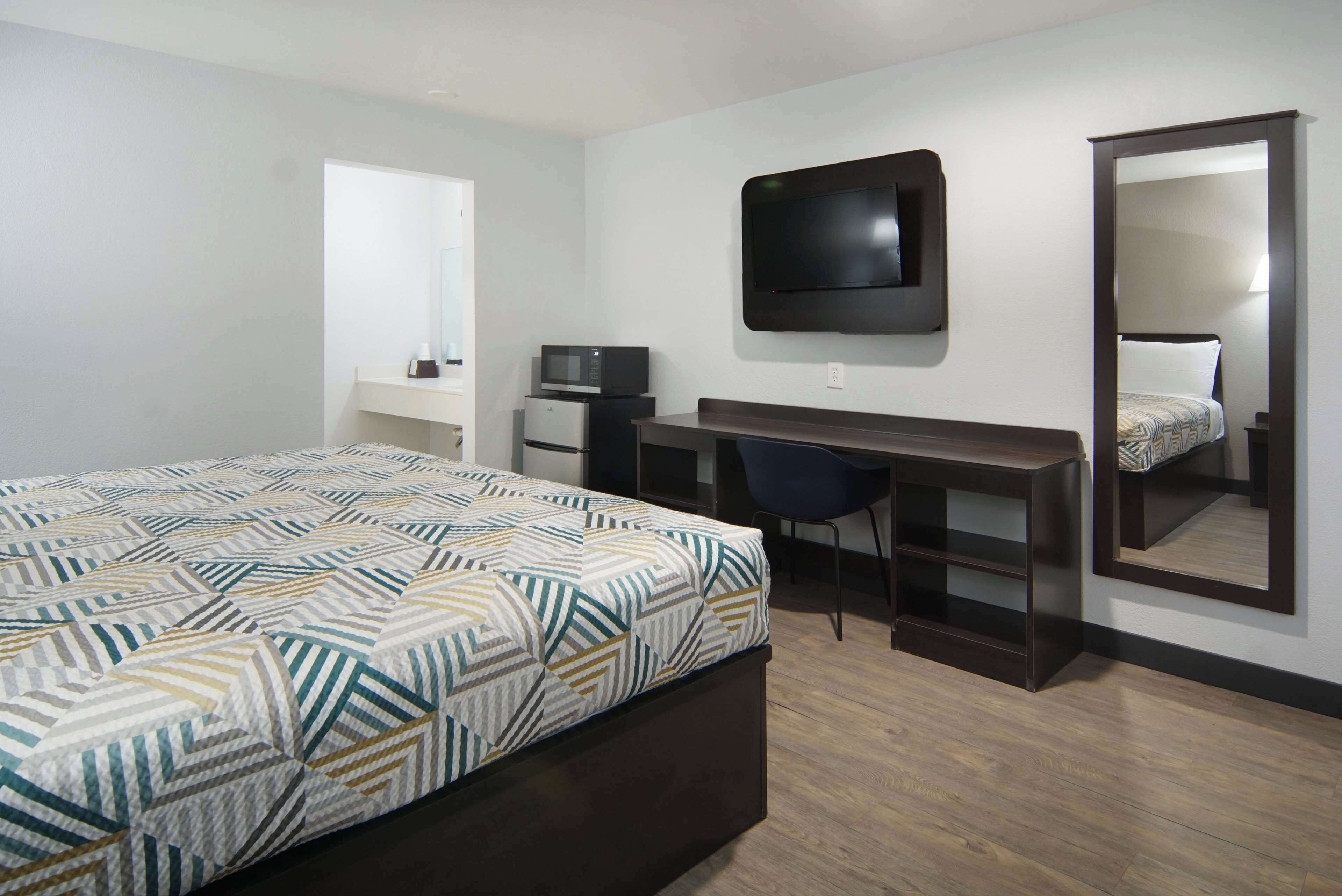 Quarto de hotel com uma cama, escrivaninha, cadeira, televisão, micro-ondas e um espelho grande.