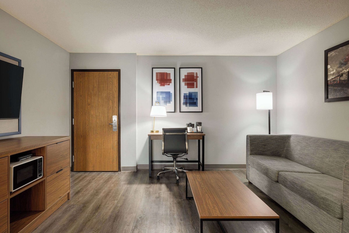 Una habitación de hotel con un escritorio de madera, una silla de oficina negra, un sofá gris, una mesa de centro de madera y dos obras de arte enmarcadas en la pared.