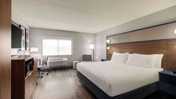A hotel room with a large bed, a desk with a flat-screen TV, a chair, a bedside table, and a window with a view.