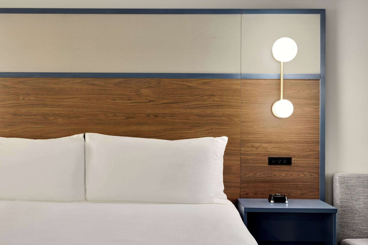Una habitación de hotel con cabecera de madera, cama con sábanas blancas, mesita de noche con lámpara y una silla.