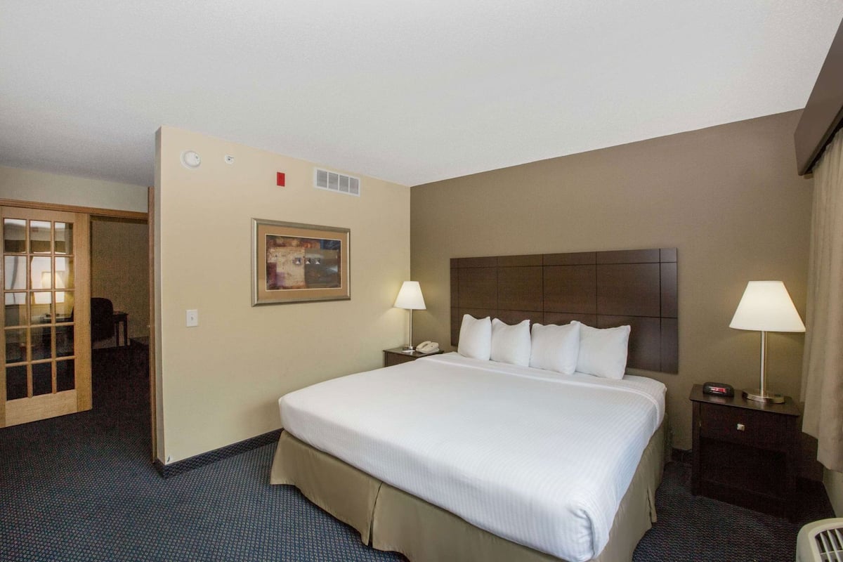 Habitación de hotel con cama, dos mesitas de noche, una lámpara de pared, un cuadro enmarcado y una puerta que da a otra habitación.