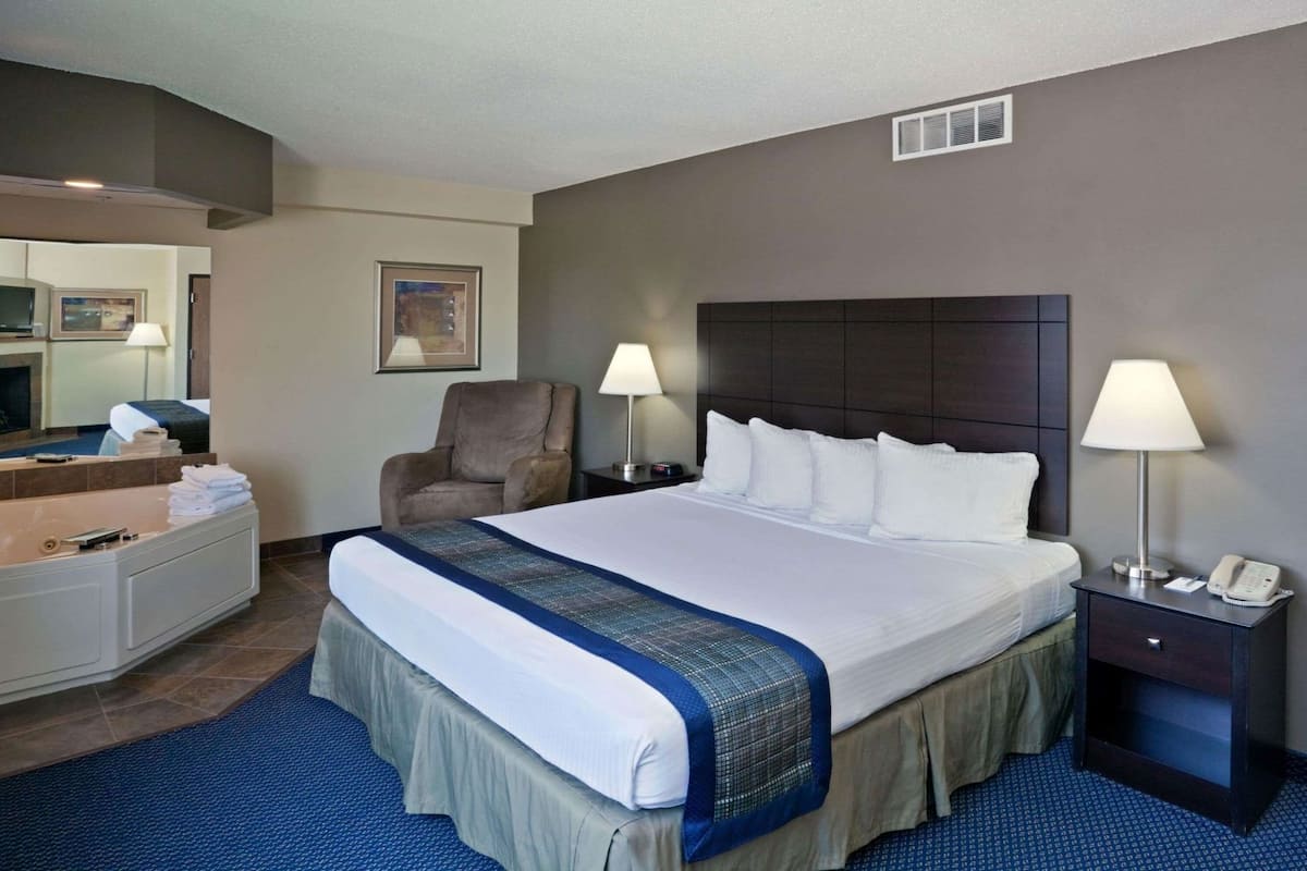 Habitación de hotel con una cama grande, dos lámparas de noche, una silla, un escritorio con lámpara, un espejo y zona de baño.