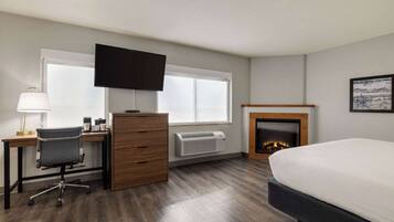 A hotel room with a bed, desk, chair, dresser, TV, fireplace, and large windows.