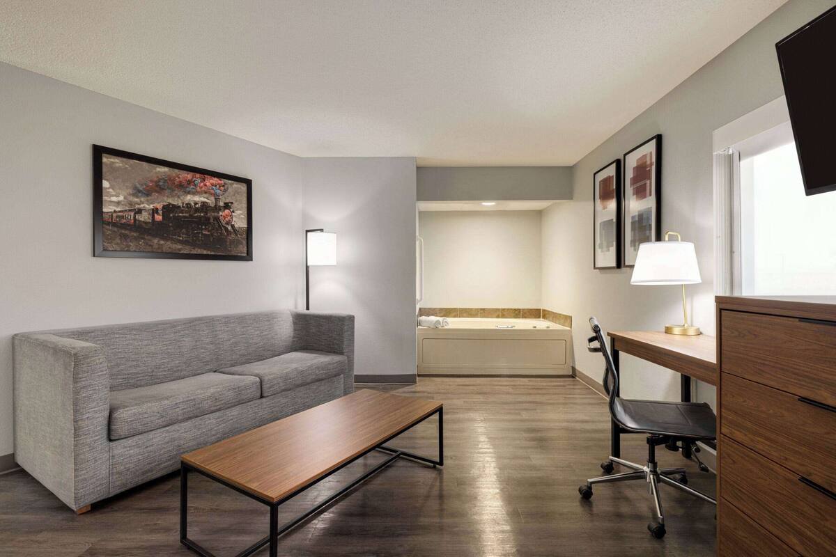 Una habitación de hotel moderna con un sofá gris, una mesa de centro de madera, un escritorio con una silla, un cuadro grande en la pared, una bañera y una ventana con televisión.
