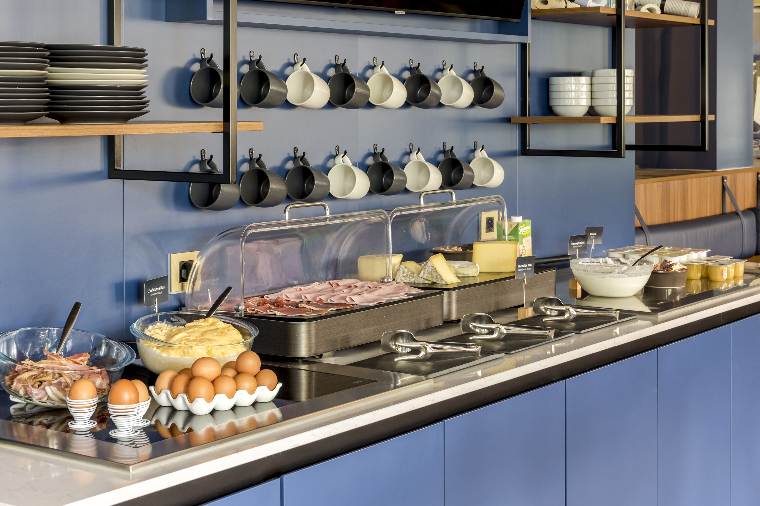 Un espace buffet proposant divers plats, notamment des œufs, de la viande et du fromage, ainsi qu’un assortiment de bols et d’assiettes.