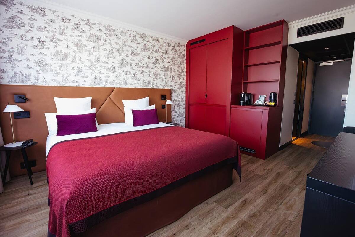 Une chambre d’hôtel avec un lit, une armoire rouge, une table de chevet et un mur orné d’un motif floral.