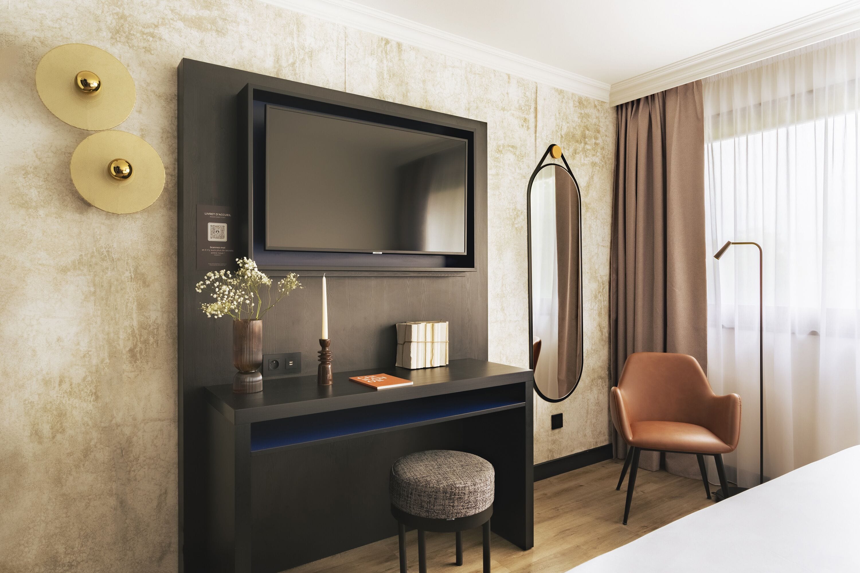 A modern hotel room with a flat-screen TV mounted on the wall, a dark console table with a vase of flowers, a small round ottoman, and a leather armchair.