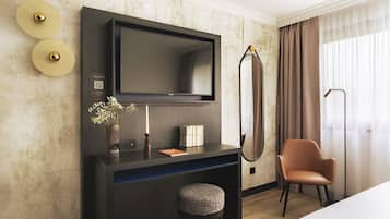 A modern hotel room with a flat-screen TV mounted on the wall, a dark console table with a vase of flowers, a small round ottoman, and a leather armchair.