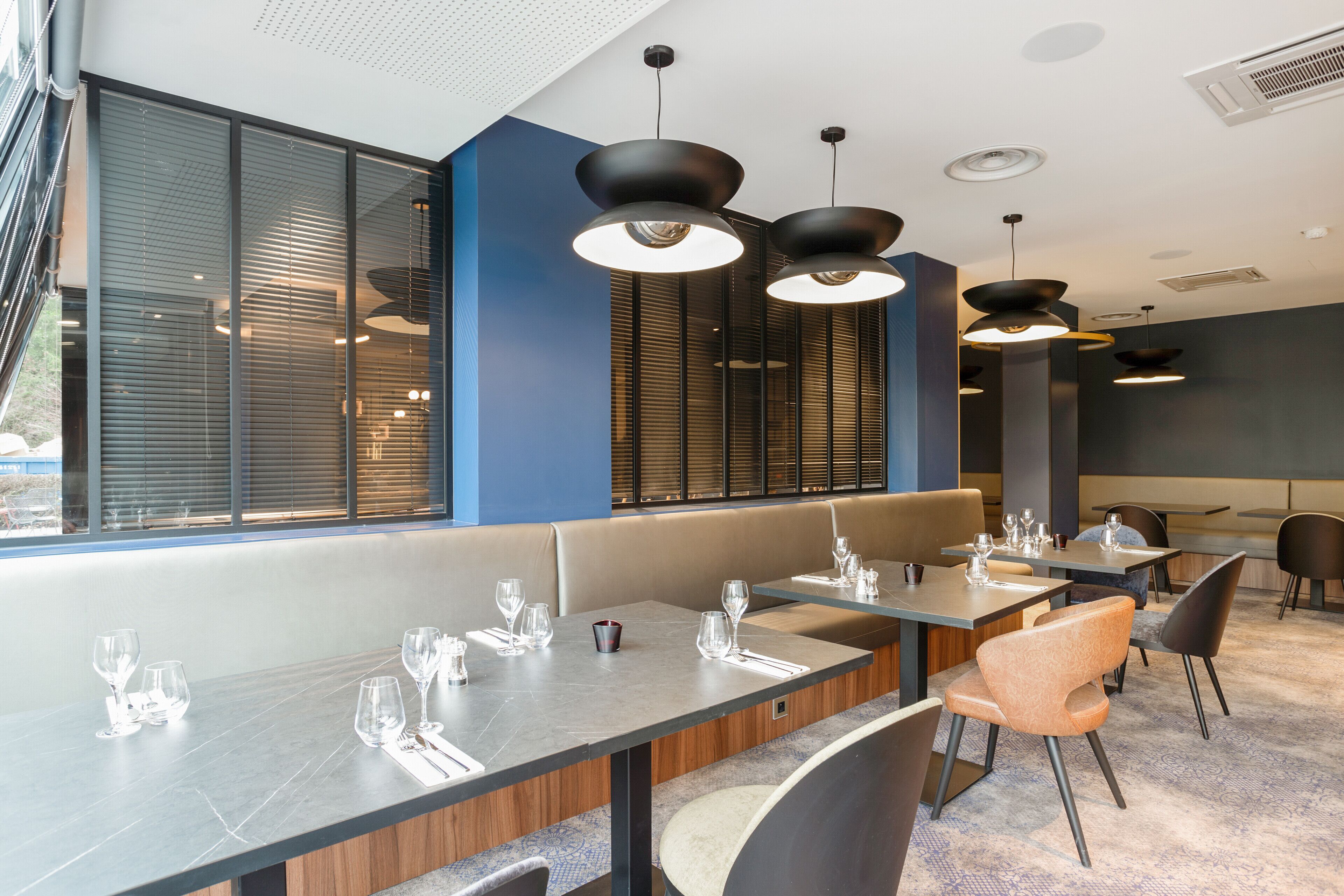 Een modern restaurant met een lange toonbank, meerdere tafels en een groot raam met zonwering.