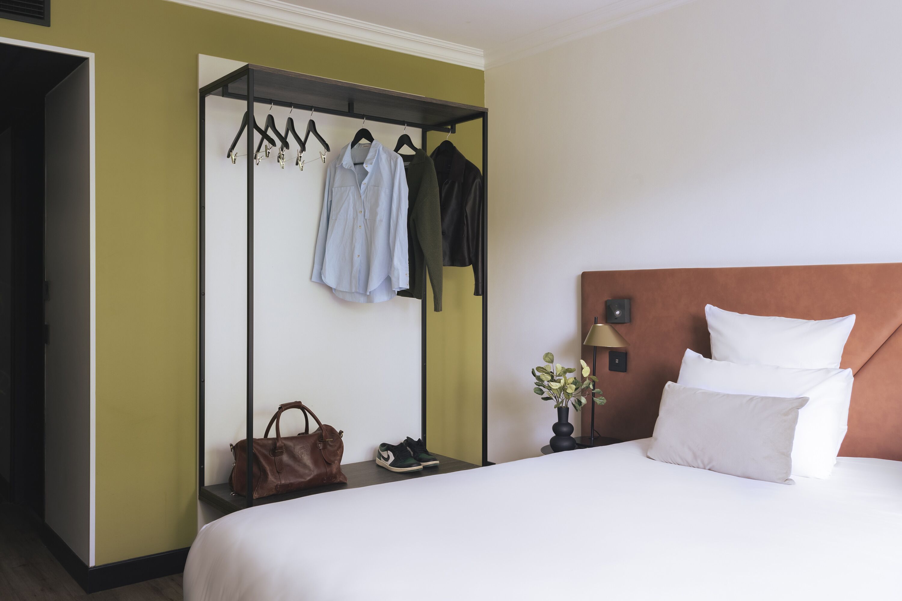 Een hotelkamer met een bed, nachtkastje, wandkapstok met hangers, handtas en een paar schoenen.