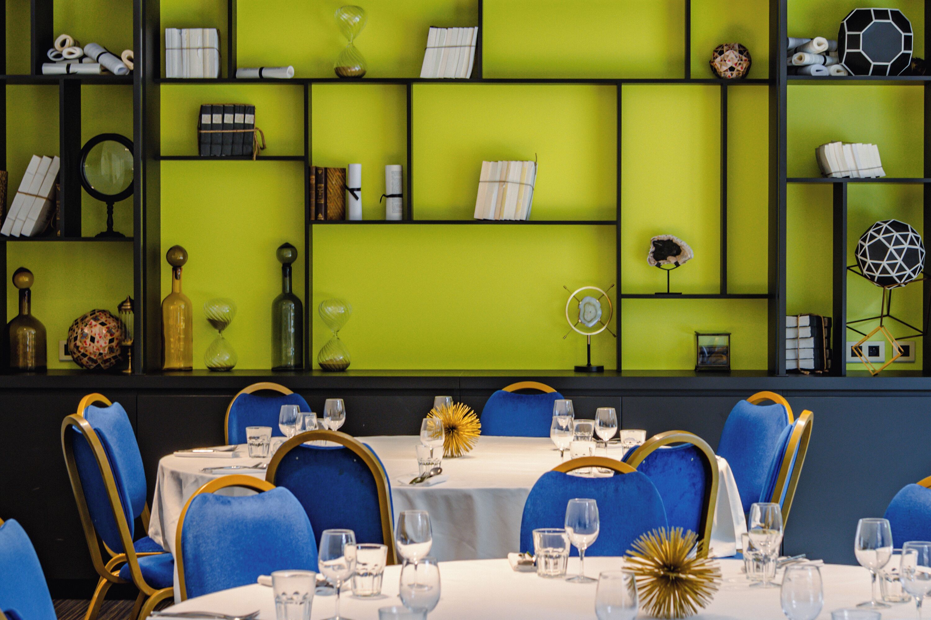 Un comedor con mesas dispuestas para el servicio, sillas con detalles dorados y estantes con diversos objetos decorativos.