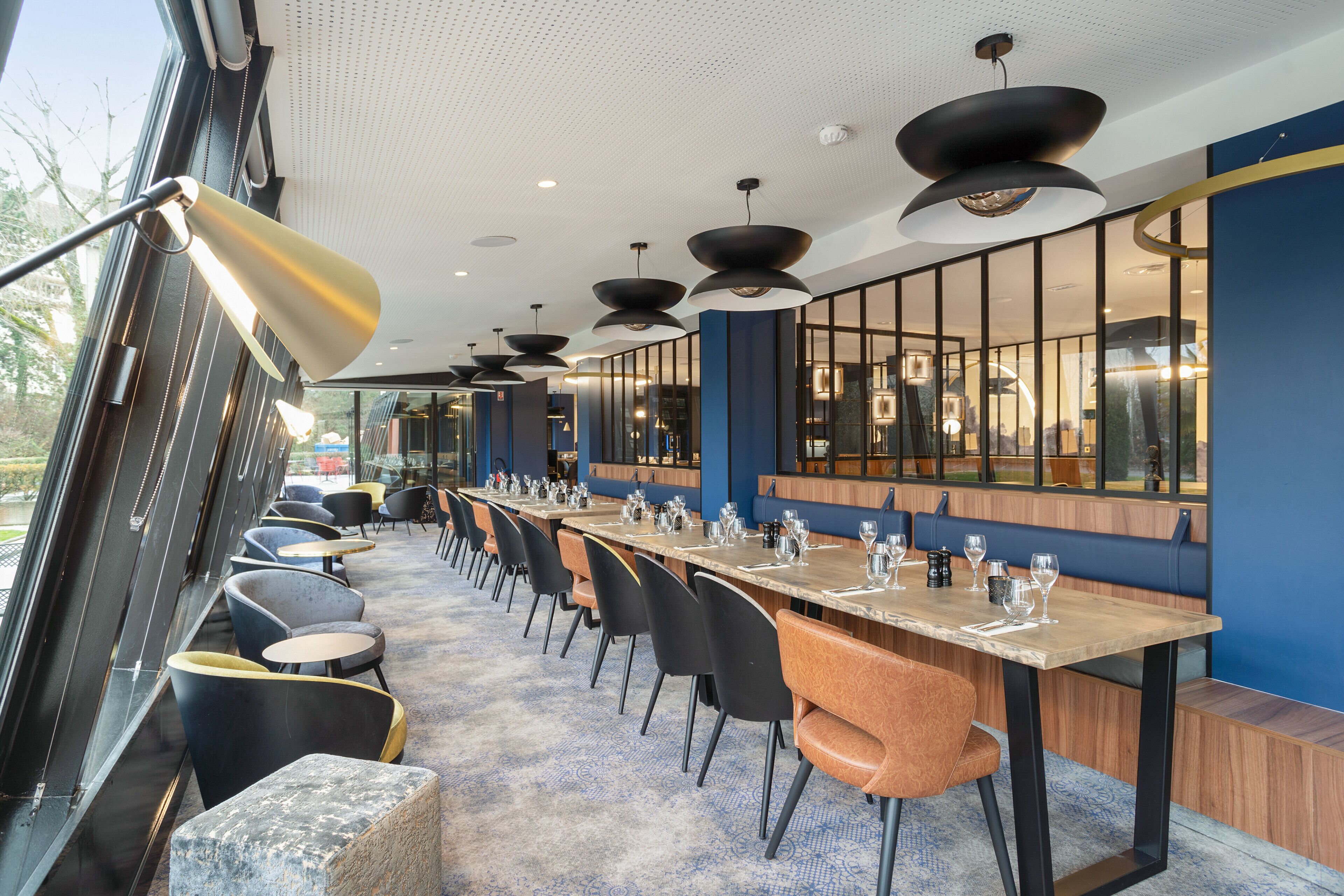 Un restaurant moderne avec une longue table, des chaises noires et orange, et de grandes fenêtres.