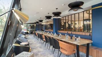 Un restaurant moderne avec une longue table, des chaises noires et orange, et de grandes fenêtres.