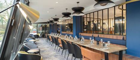 A modern restaurant with a long table, black and orange chairs, and large windows.