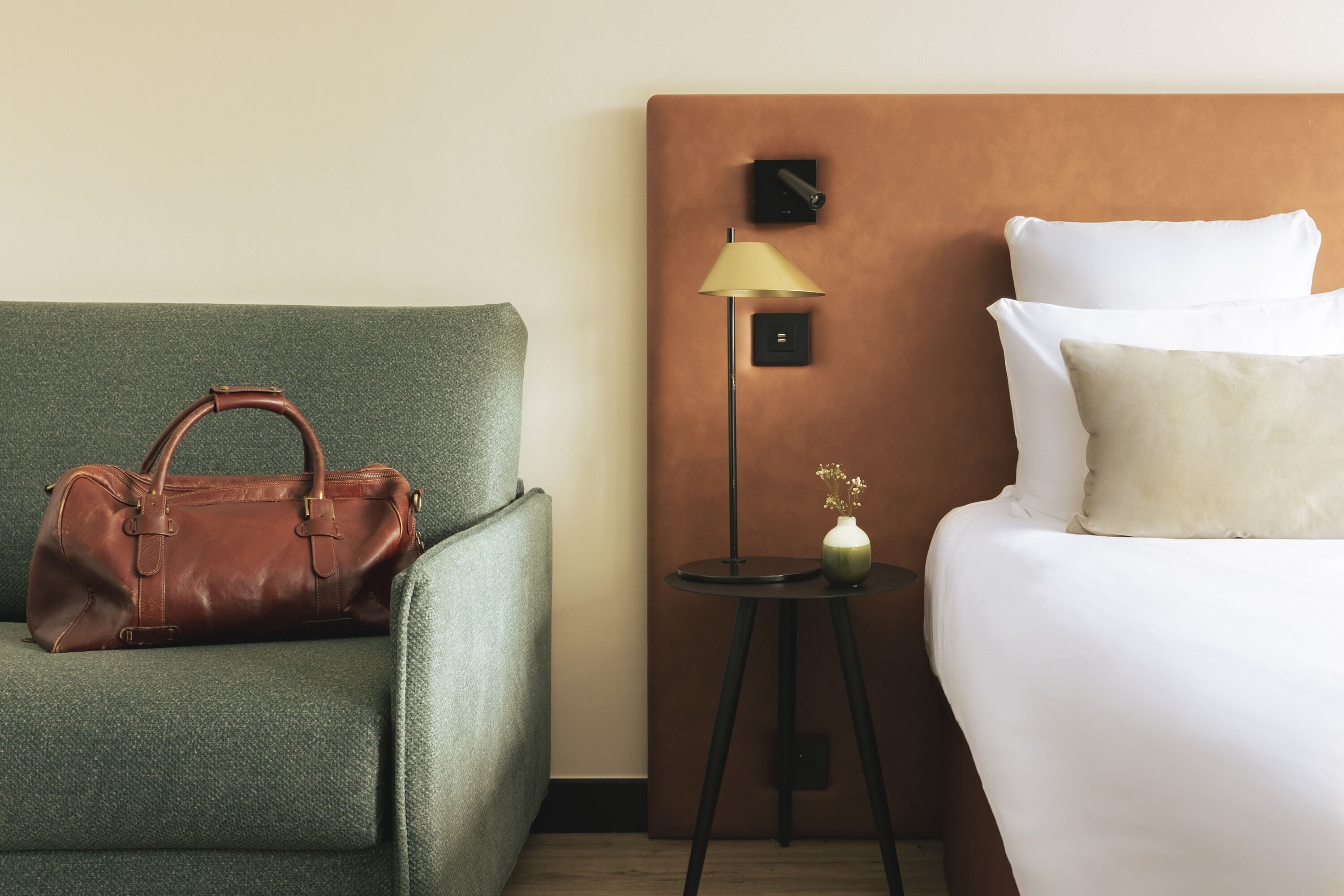 Una habitación de hotel con un sillón verde, un bolso de cuero marrón, una mesita de noche con lámpara y una cama con ropa blanca de cama.